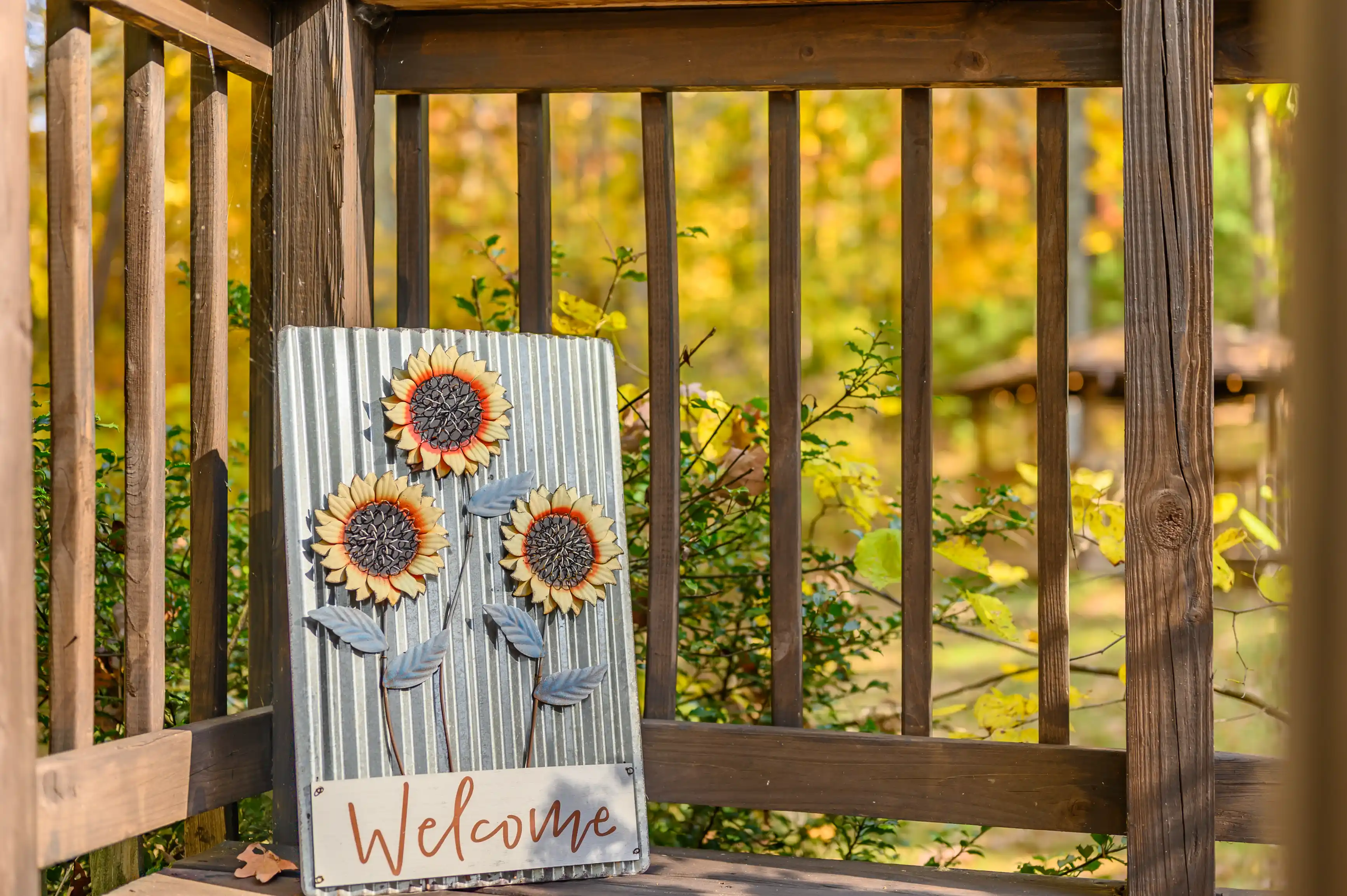 Decorative welcome sign with sunflower design on a wooden porch with autumn leaves in the background.