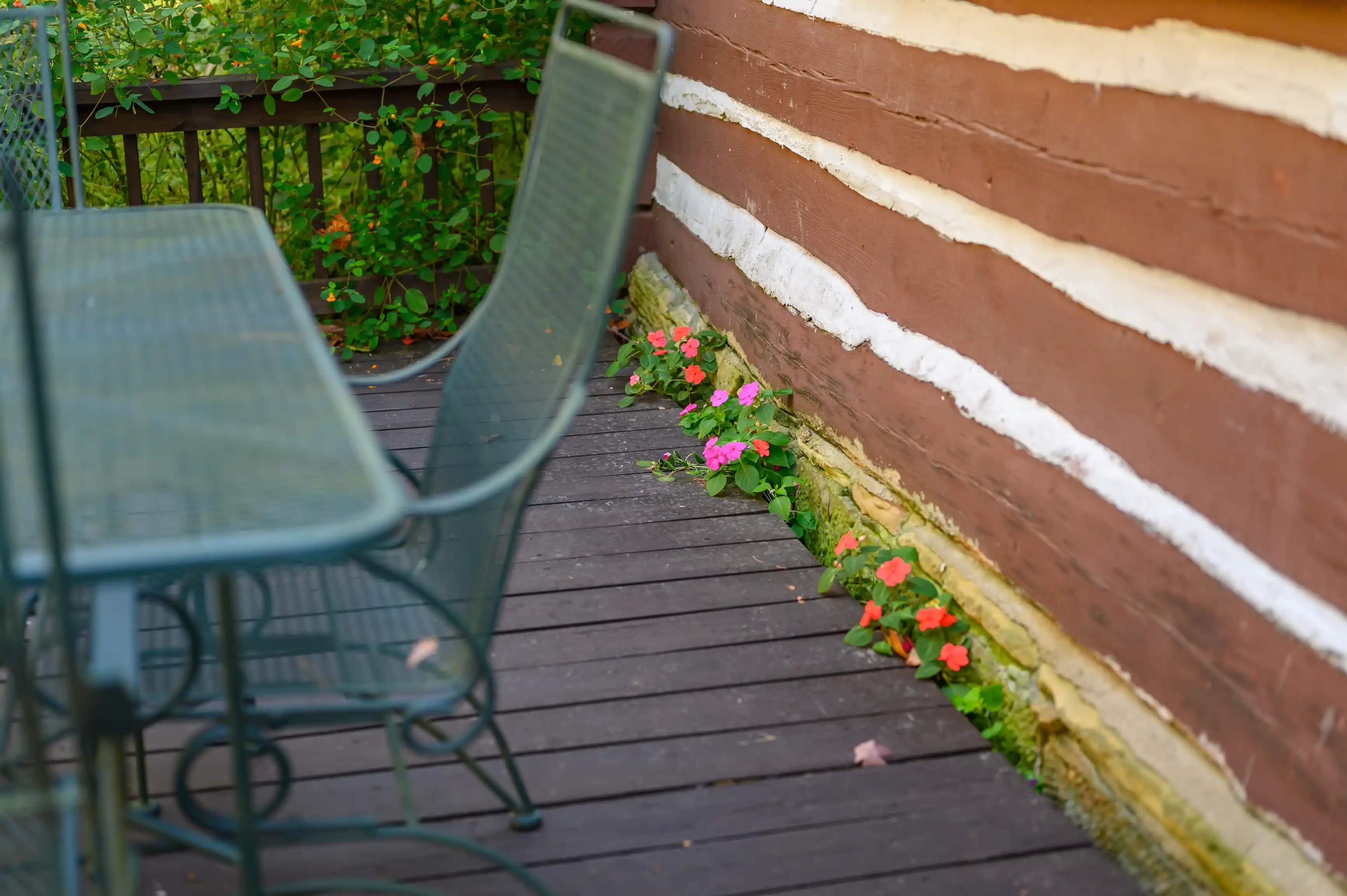 Outdoor patio area with a metal chair and table set on a wooden deck, adjacent to a house with brick siding, and some pink flowers sprouting between the planks.
