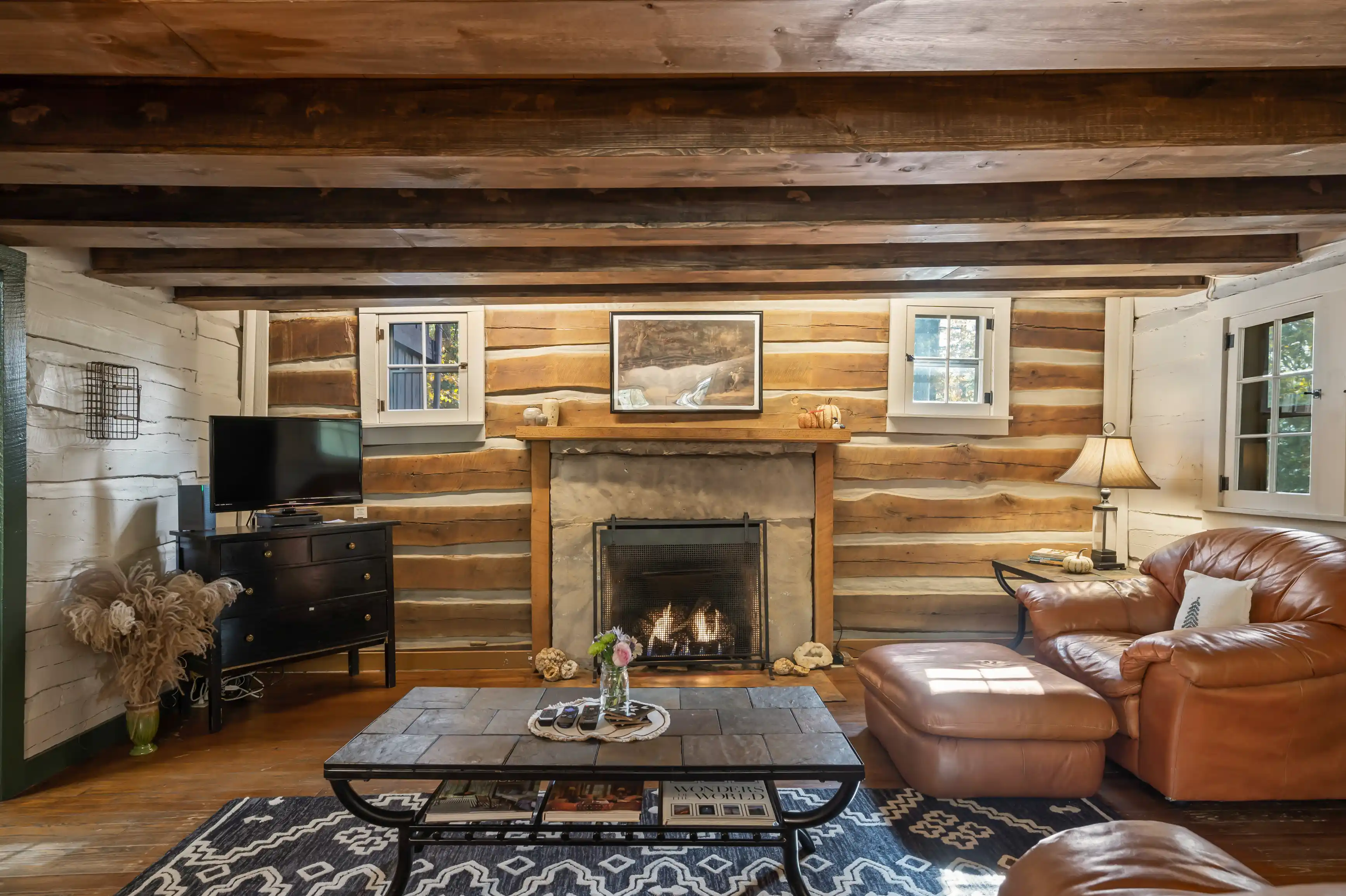 Cozy cabin living room interior with a lit fireplace, leather sofas, wooden walls, a Persian rug, and a flat-screen TV.