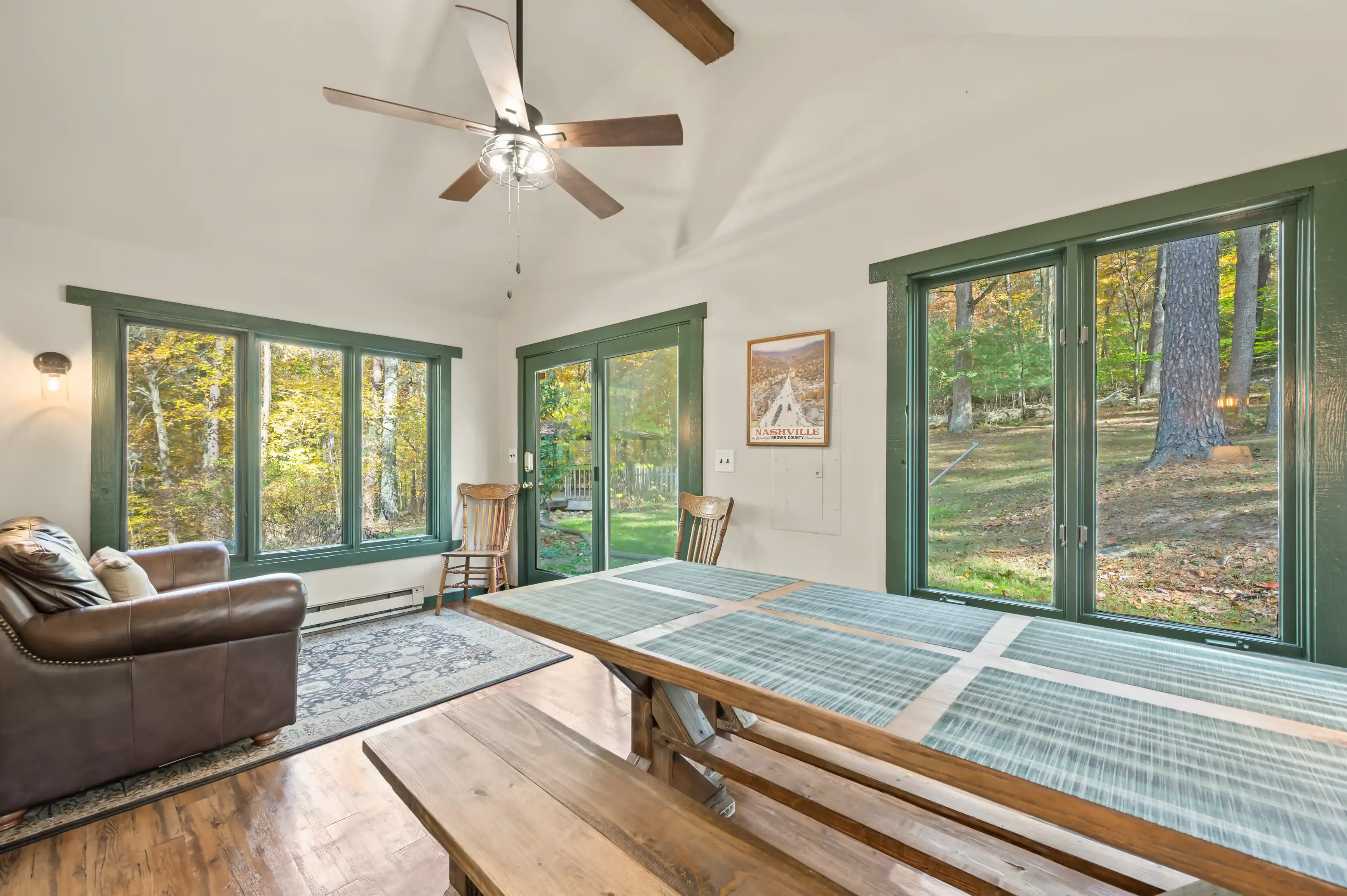 Bright and airy sunroom with large windows overlooking trees, furnished with a ping-pong table, a brown leather sofa, and a ceiling fan above.