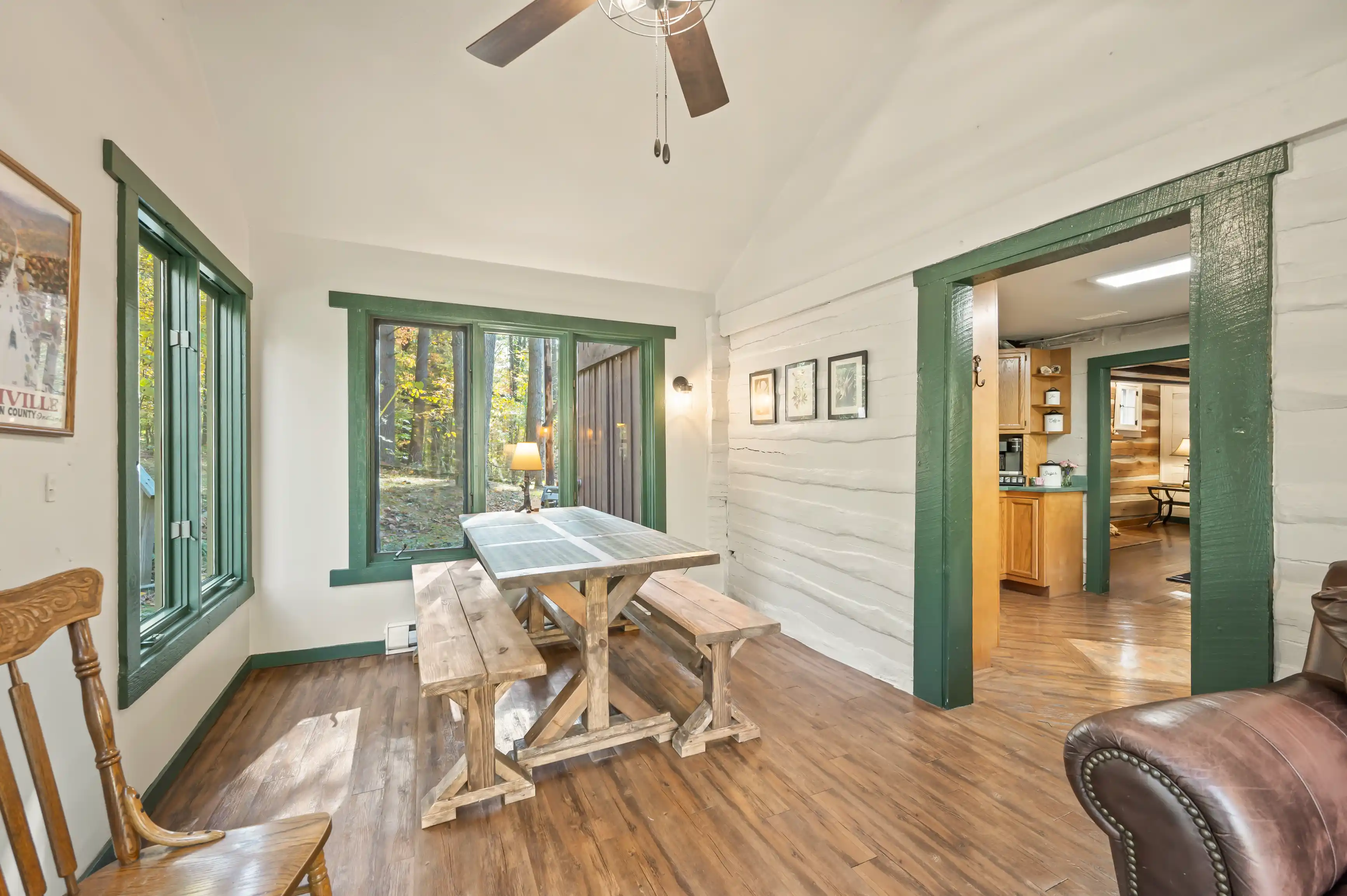 Cozy cottage-style dining area with a wooden picnic table and benches, hardwood floors, a rocking chair, and green-trimmed windows with views of nature outside.