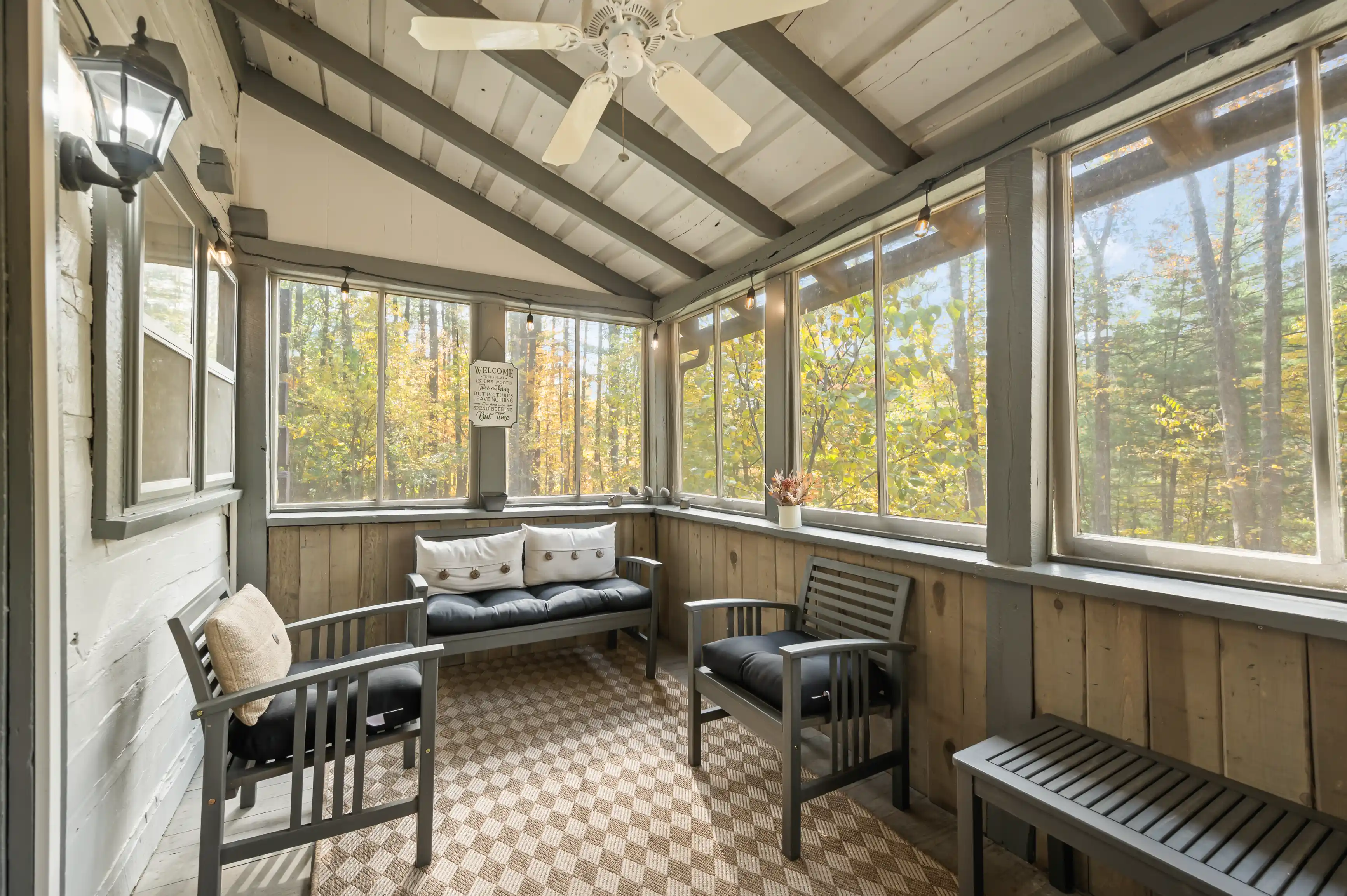 Cozy sunroom with a ceiling fan, large windows with fall foliage views, and seating furniture.