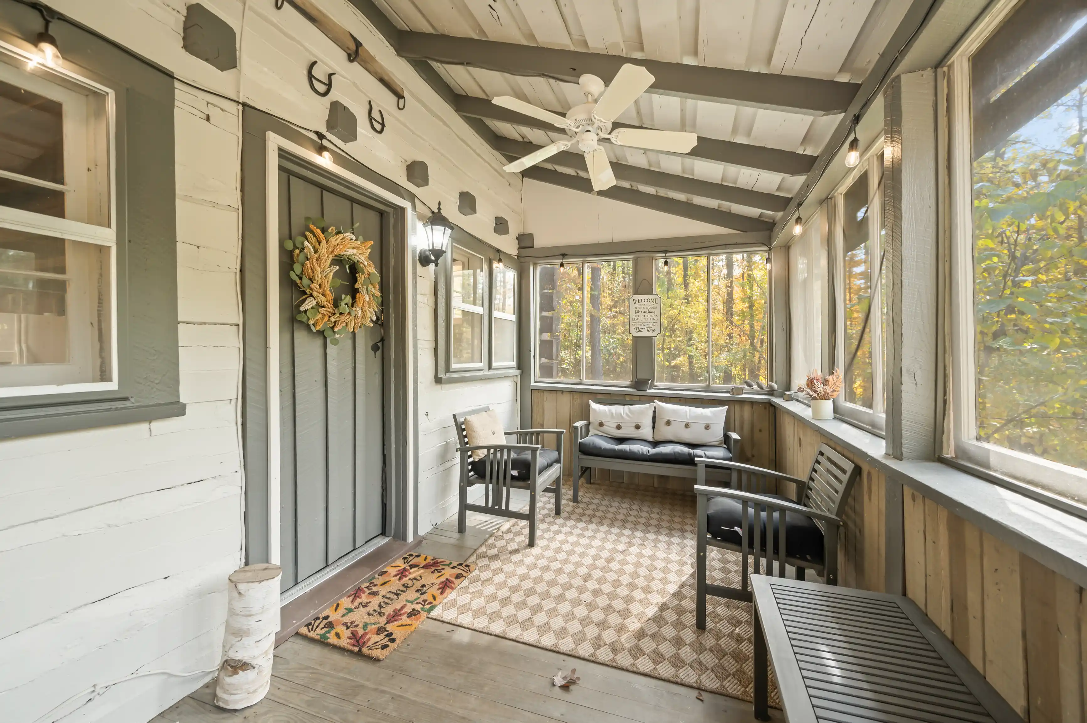 Cozy screened porch with wicker furniture, ceiling fan, and autumn wreath on the door, surrounded by trees visible through the screens.