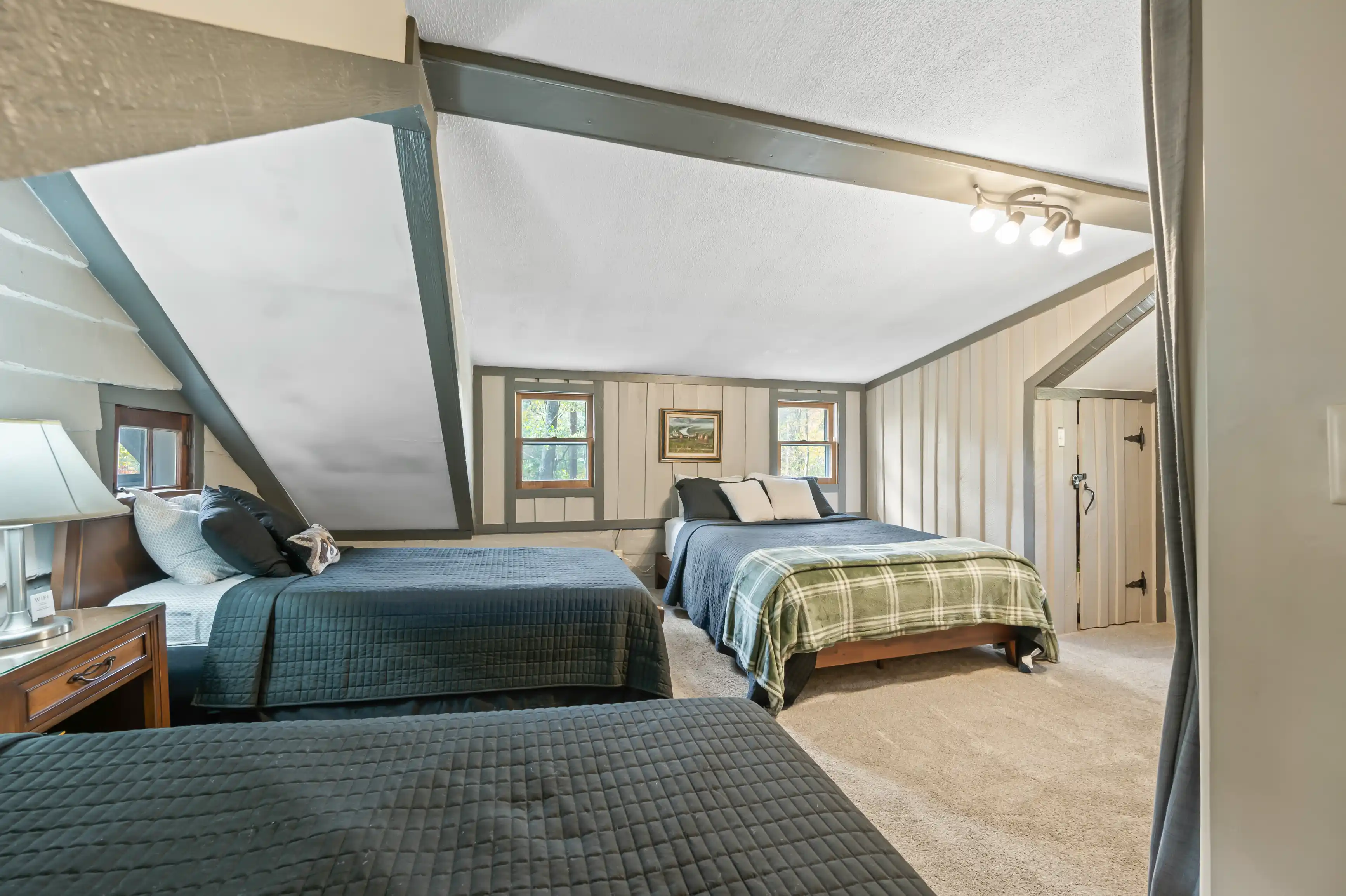 Bright and cozy attic bedroom interior with sloped ceilings, two beds with quilts, nightstands with lamps, framed artwork, and a carpeted floor.