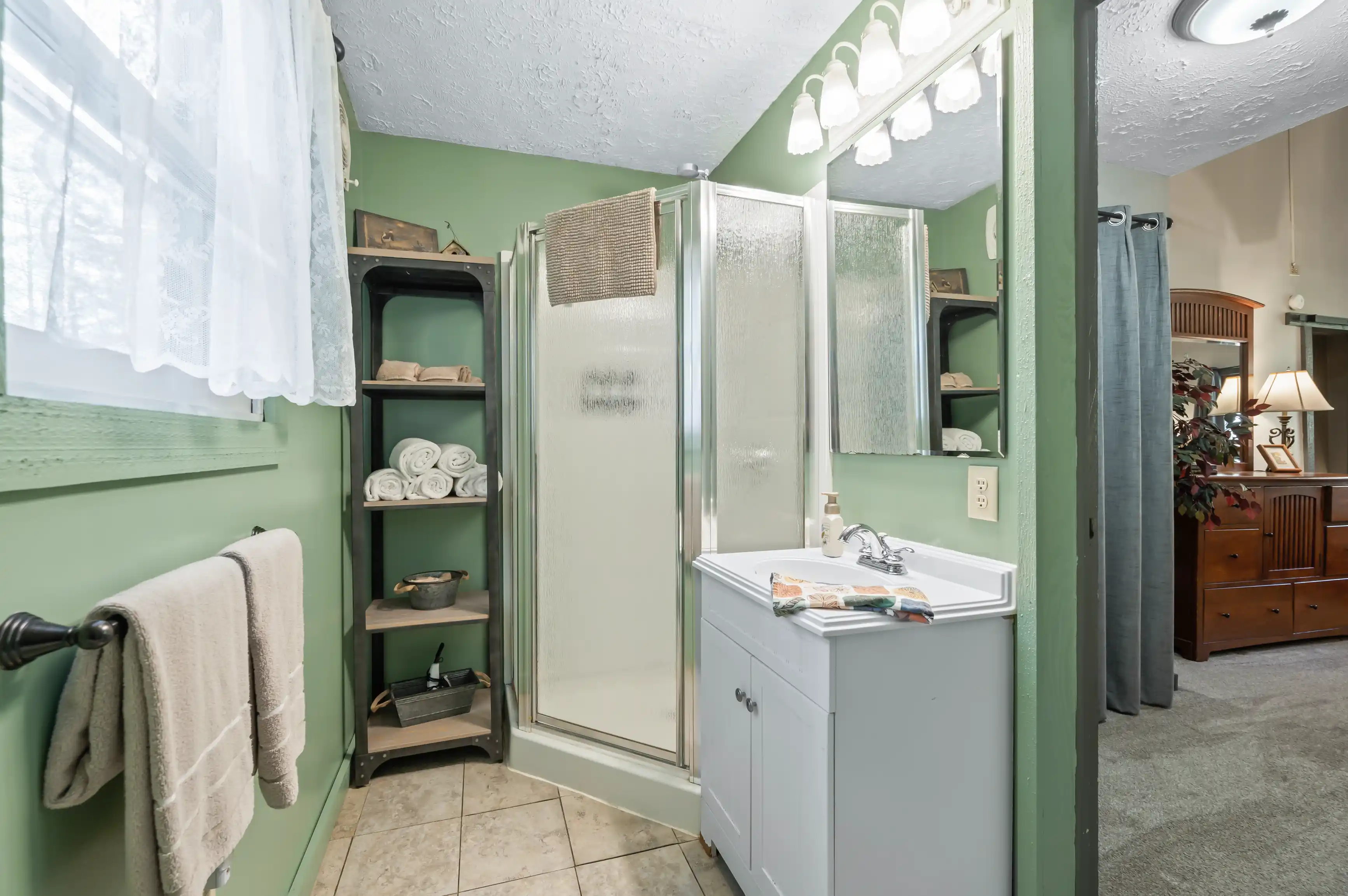 Bathroom interior with light green walls, shower stall, vanity with sink, mirror with overhead lights, towel rack, and adjacent room visible in the background.