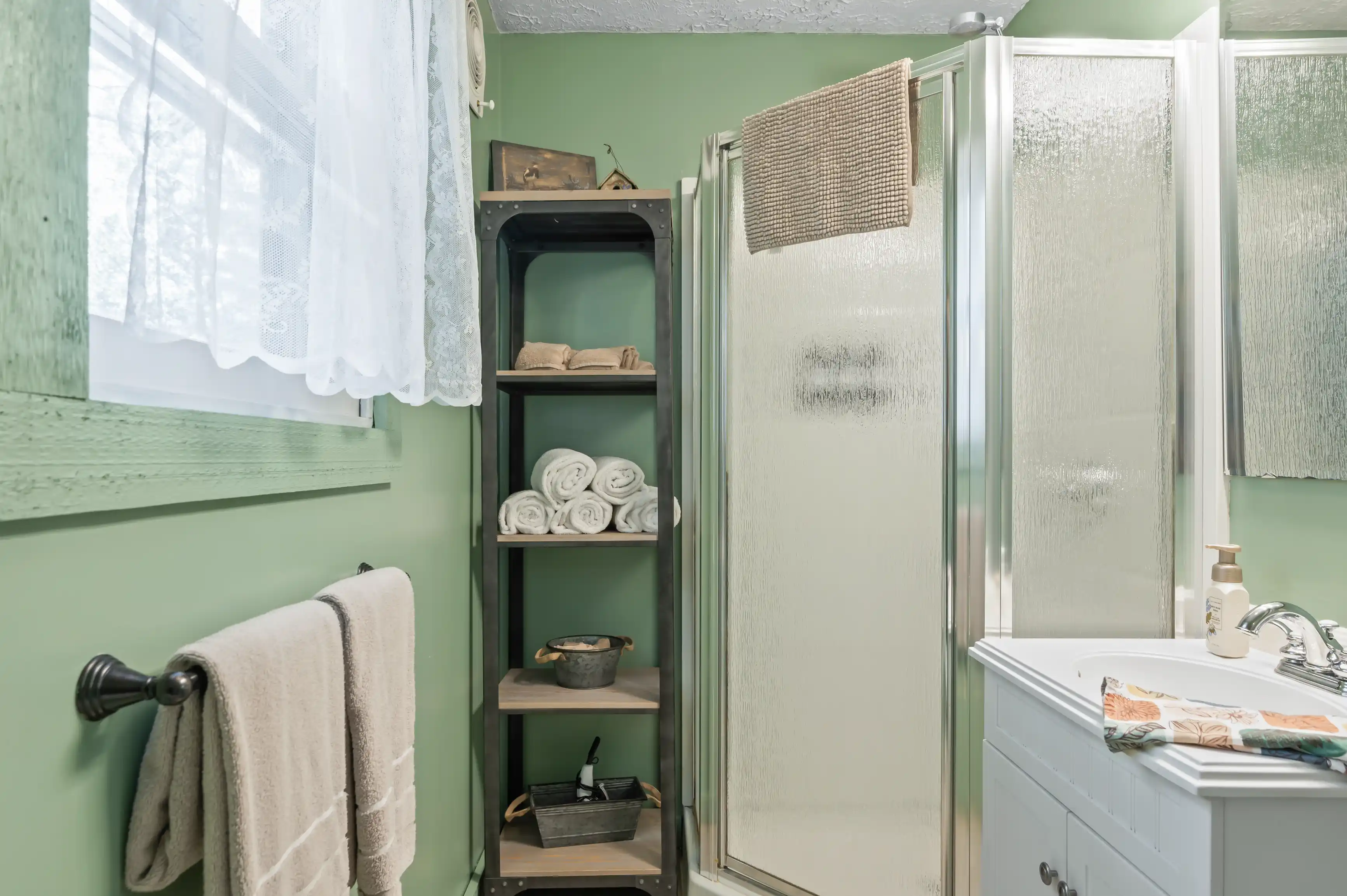 Bright bathroom with green walls, white fixtures, shower stall, and a wooden shelving unit with towels.