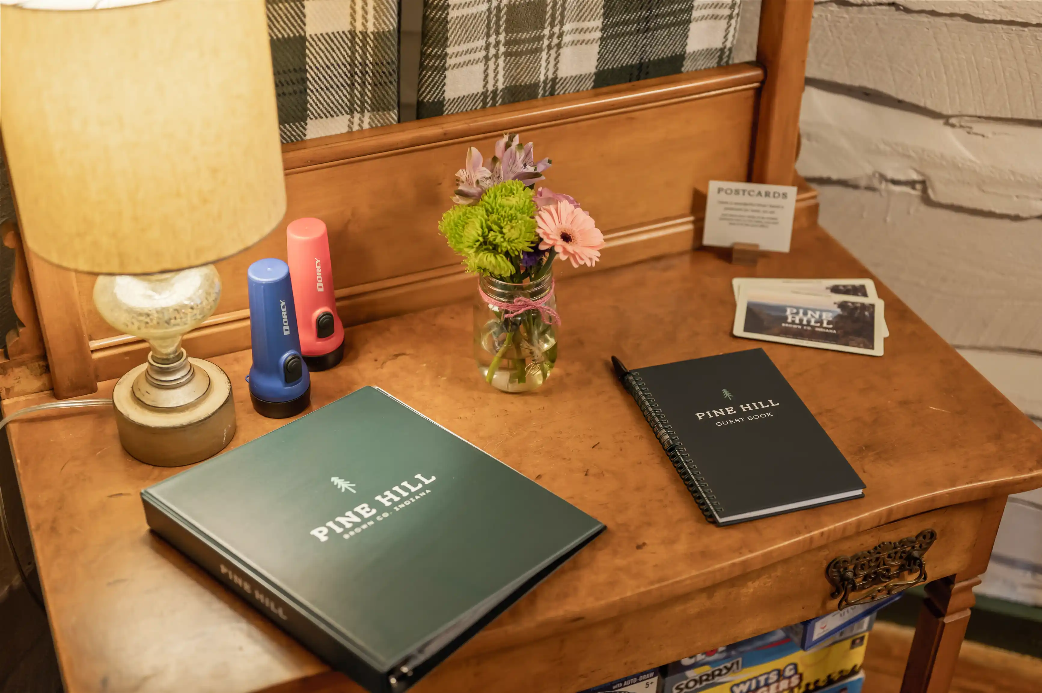 Cozy workspace with a lamp, a small vase of flowers, a green book titled "My Wellness Journal," a black book titled "Work Plan," and some pens on a wooden desk.