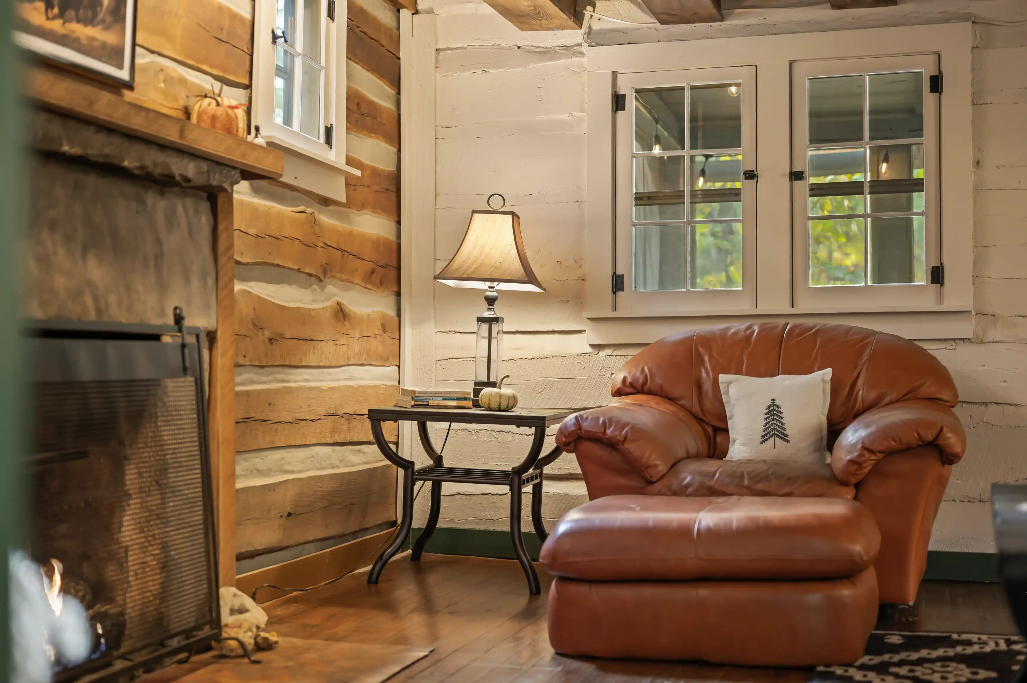 Cozy cabin interior with a plush leather armchair, wooden side table with a lamp, and part of a fireplace, against a rustic wooden wall with a window.