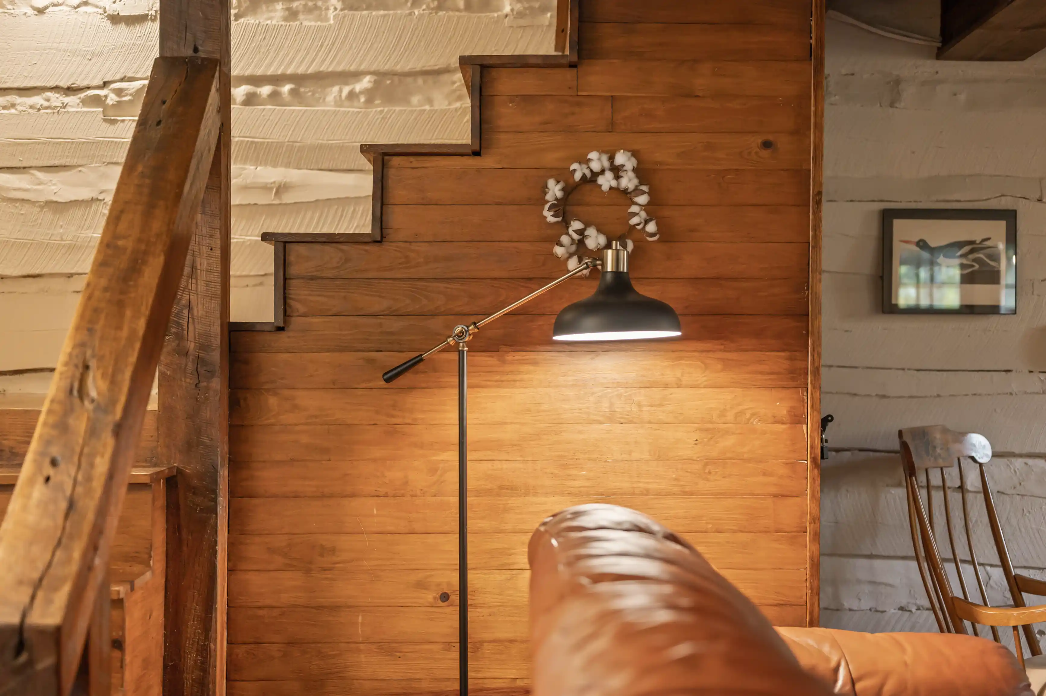 Cozy interior corner with leather sofa, wooden staircase, floor lamp, and a small bouquet on the wall.