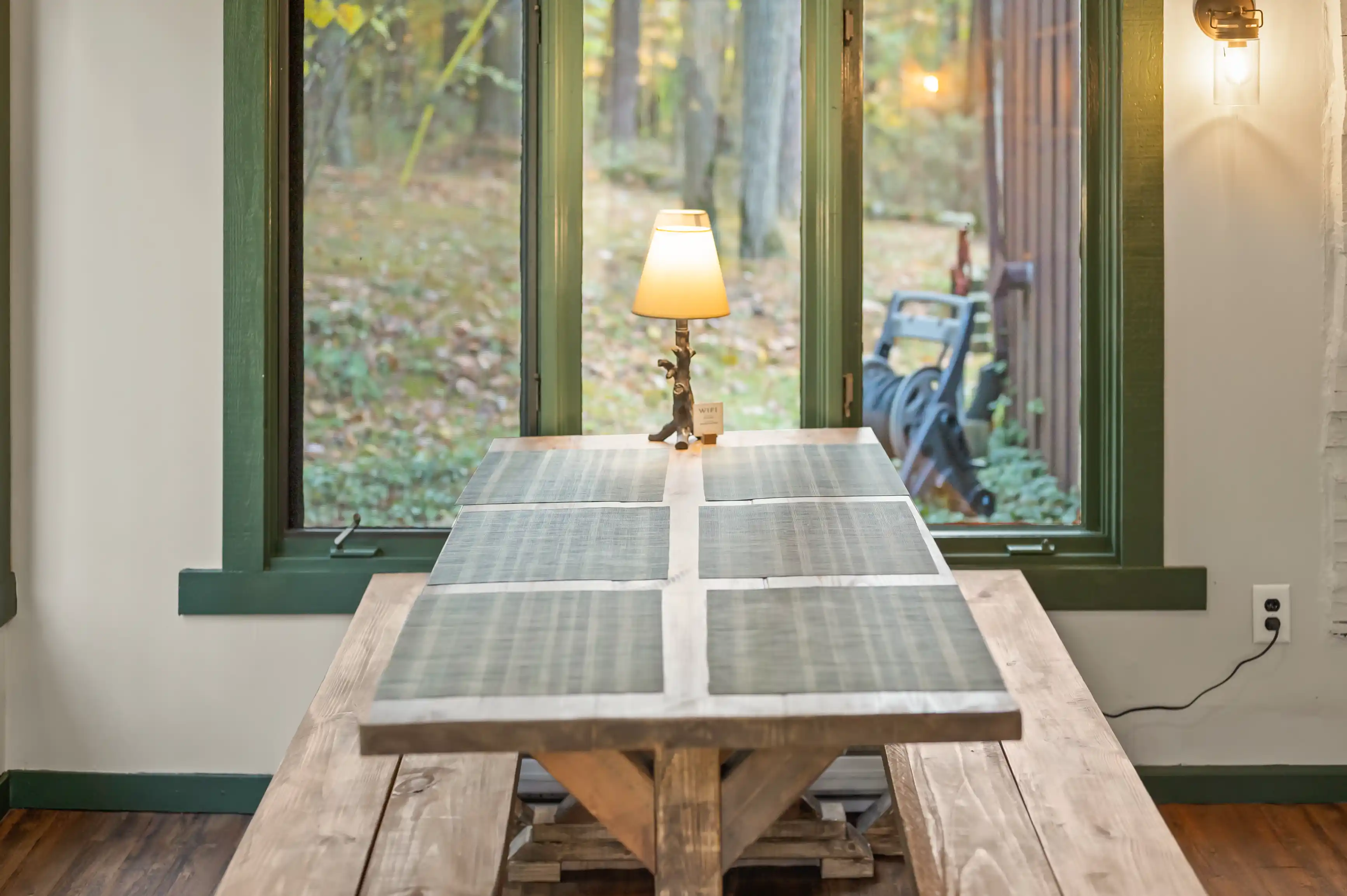 Cozy interior view of a rustic wooden table with an open book and a lamp, by a window overlooking a forest.