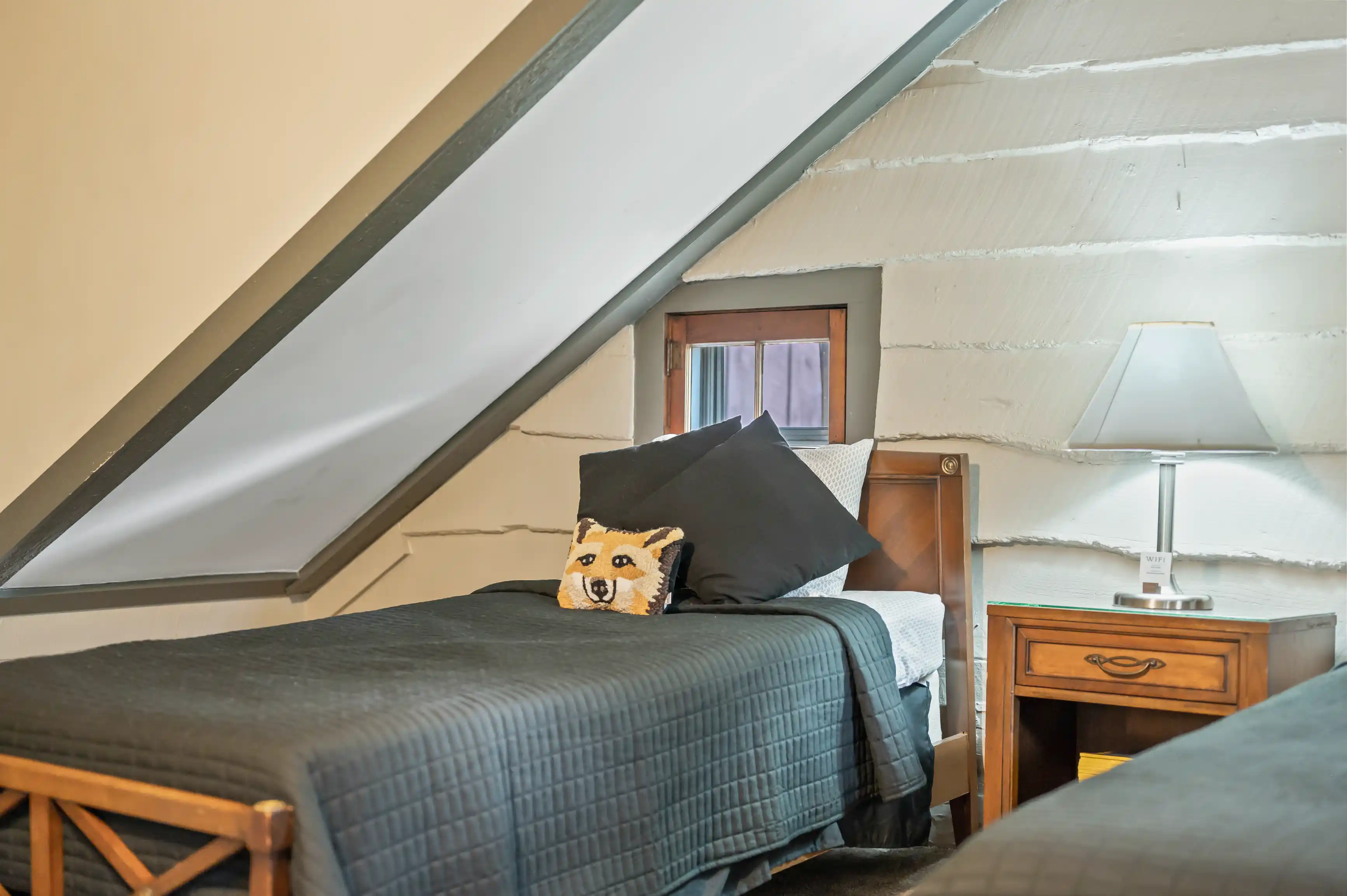 Cozy attic bedroom with a sloped ceiling, a comfortable bed with a grey blanket, and a bedside table with a lamp.