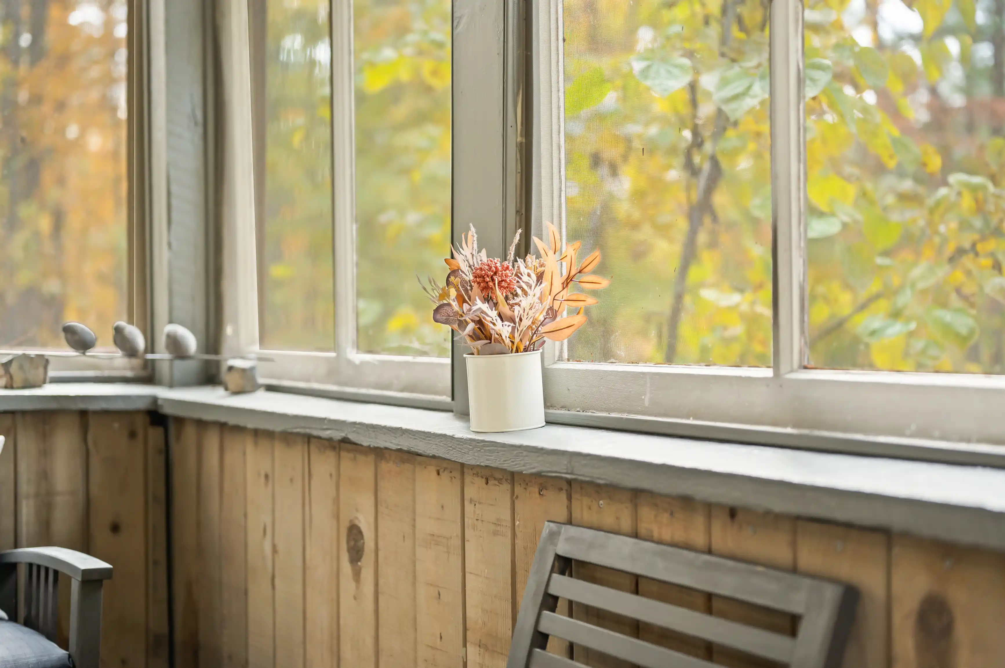 A cozy window nook with a wooden sill, a white vase with dried flowers, and a view of autumn foliage outside.