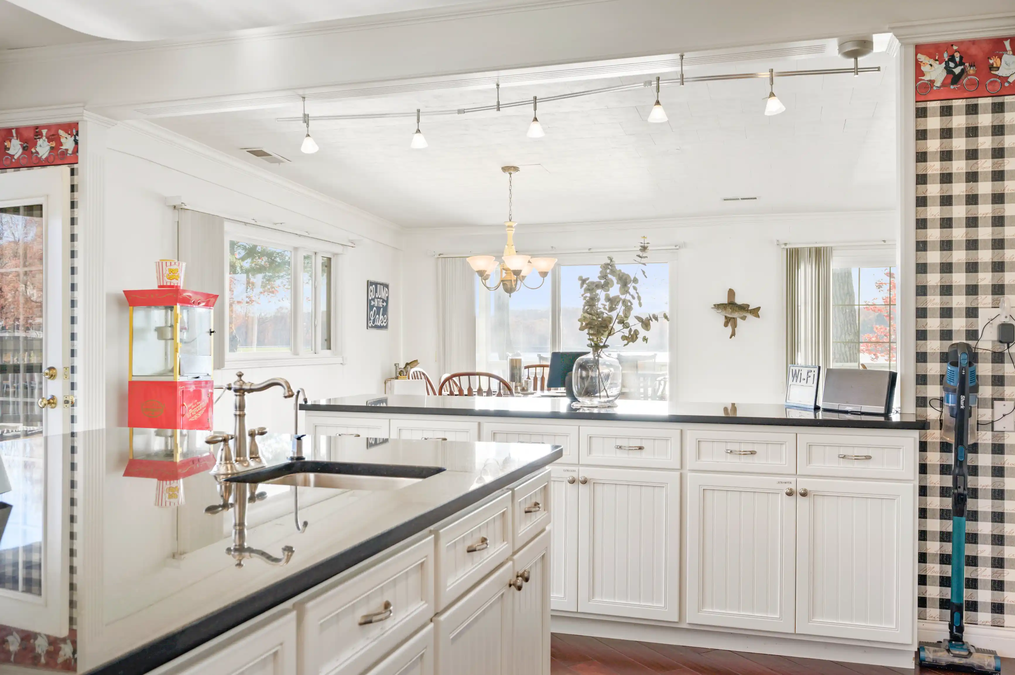 Bright kitchen with white cabinets, black countertops, a vintage popcorn machine, plaid wall decor, and a chandelier over an island.