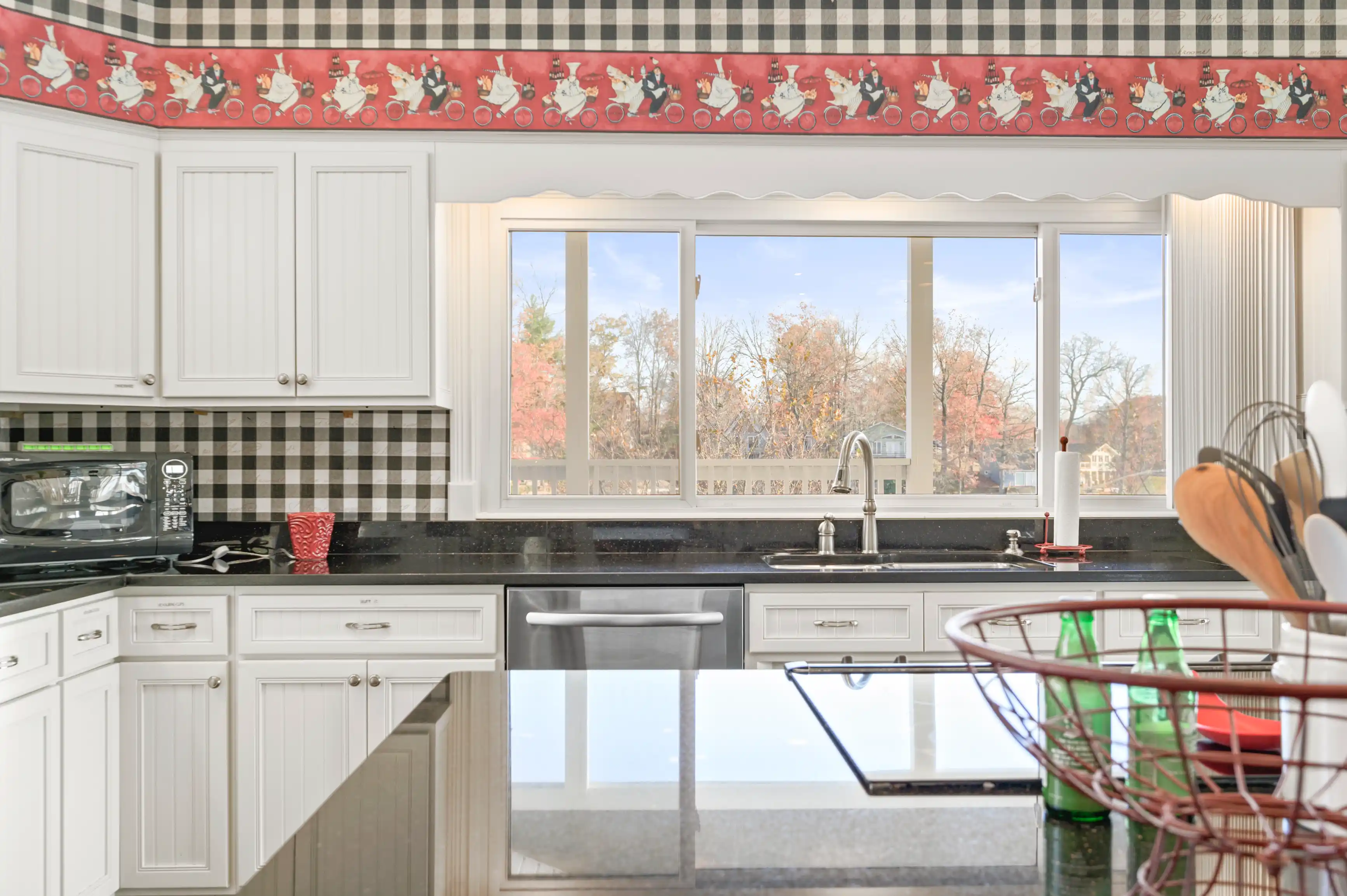 Bright kitchen interior with white cabinetry, black countertops, checkered backsplash, and a picturesque window view with autumn foliage.