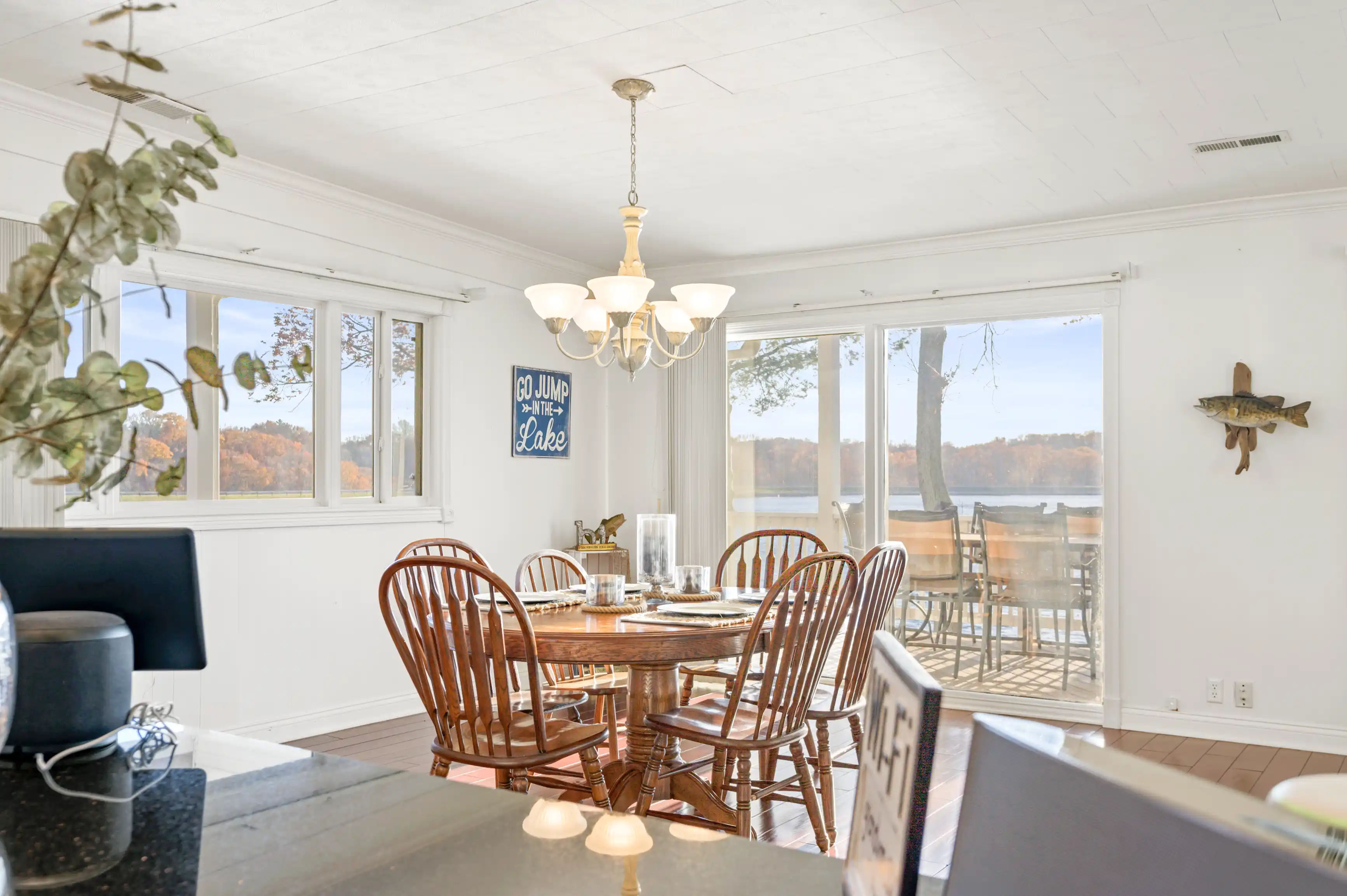 Bright dining room interior with wooden table set, decorative sign on the wall, and scenic lake view through large windows.