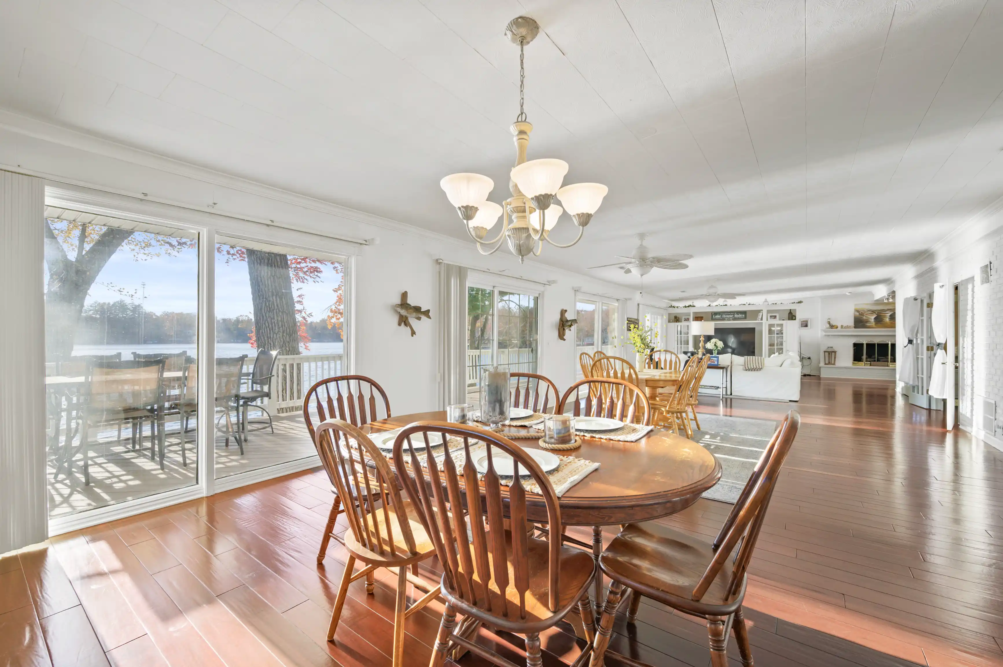 Bright and spacious dining room with a wooden oval table and chairs, opening onto a sunny deck overlooking a lake through sliding glass doors.