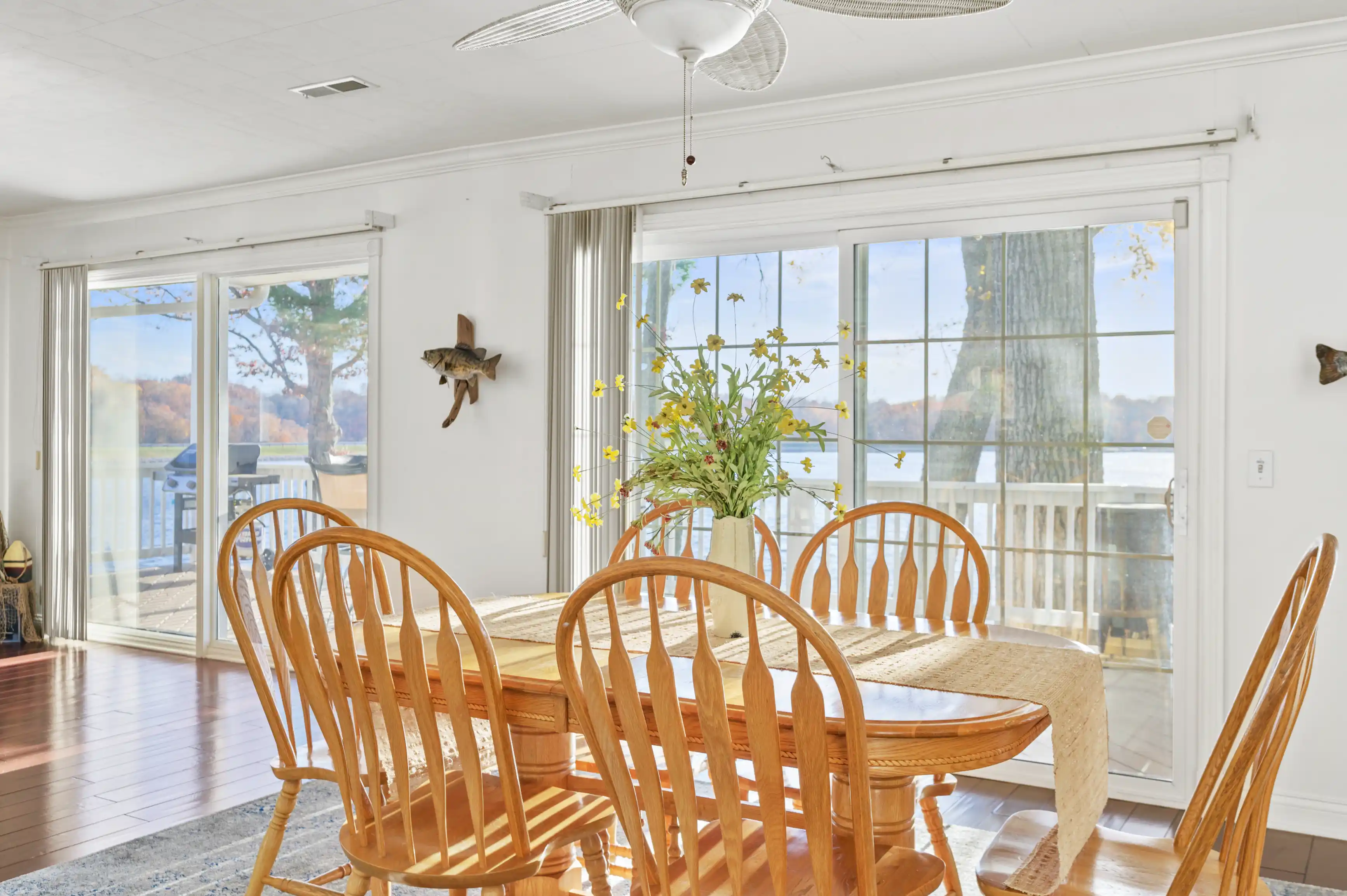 Bright and airy dining room with wooden furniture, a flower centerpiece, and lake view through sliding glass doors.