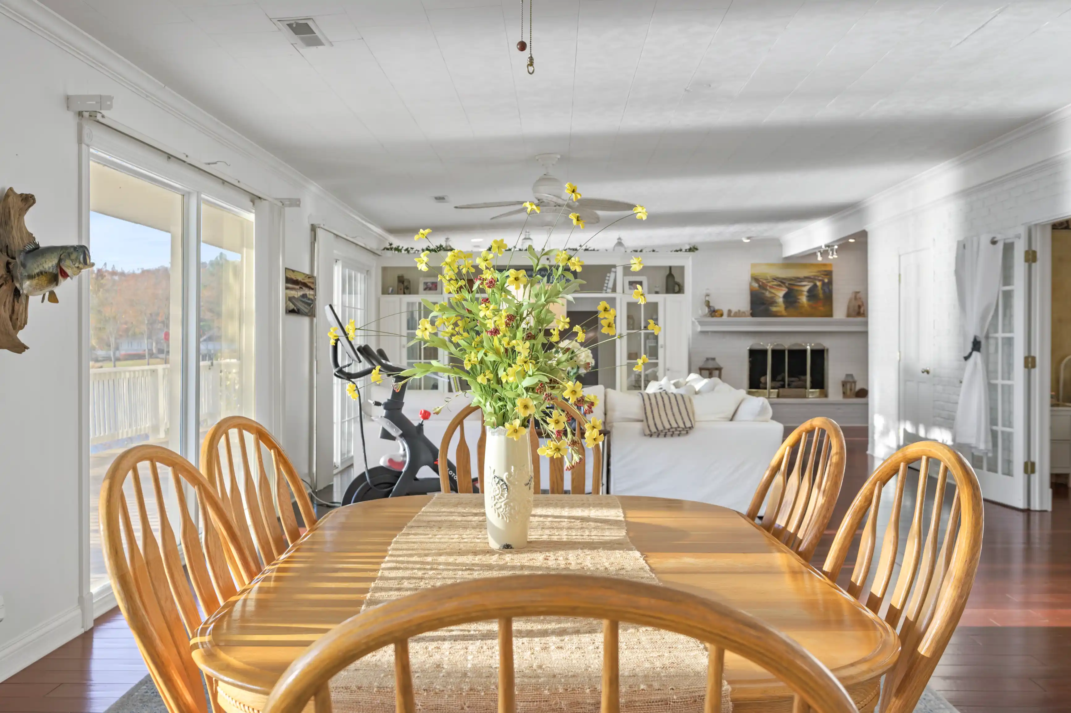 Bright and airy dining room with a wooden table and chairs, a large vase with yellow flowers, and a living area in the background with a white couch and fireplace.