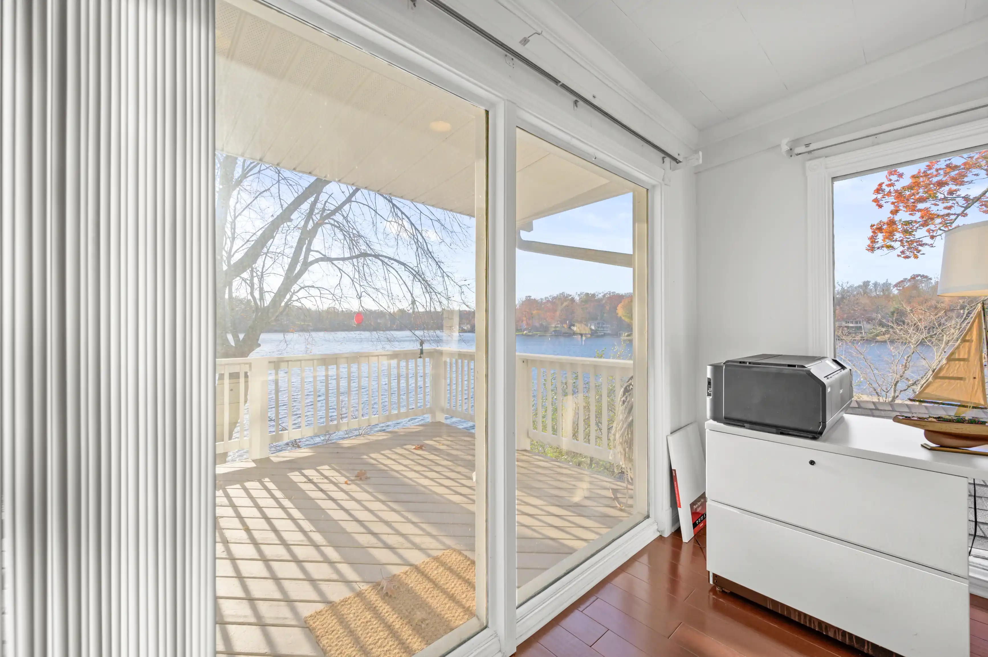Bright sunlit room with open sliding doors leading to a deck overlooking a lake, with autumn trees in the distance.