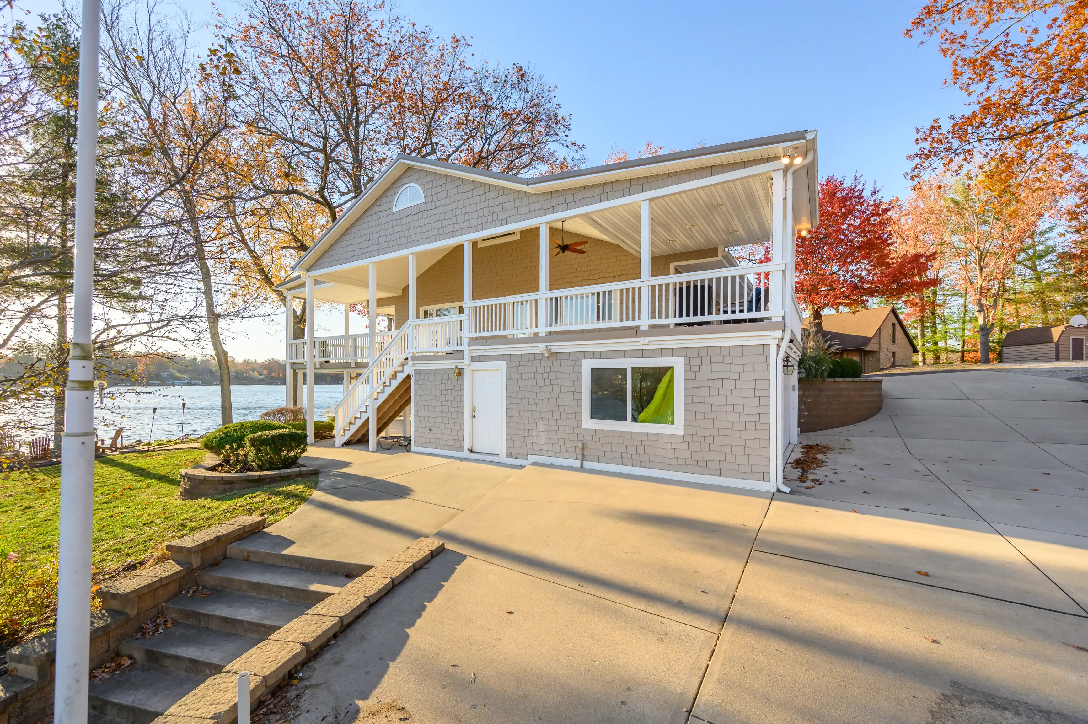 Alt: Two-story waterfront house with upper and lower white decks, autumn trees, and a clear sky.
