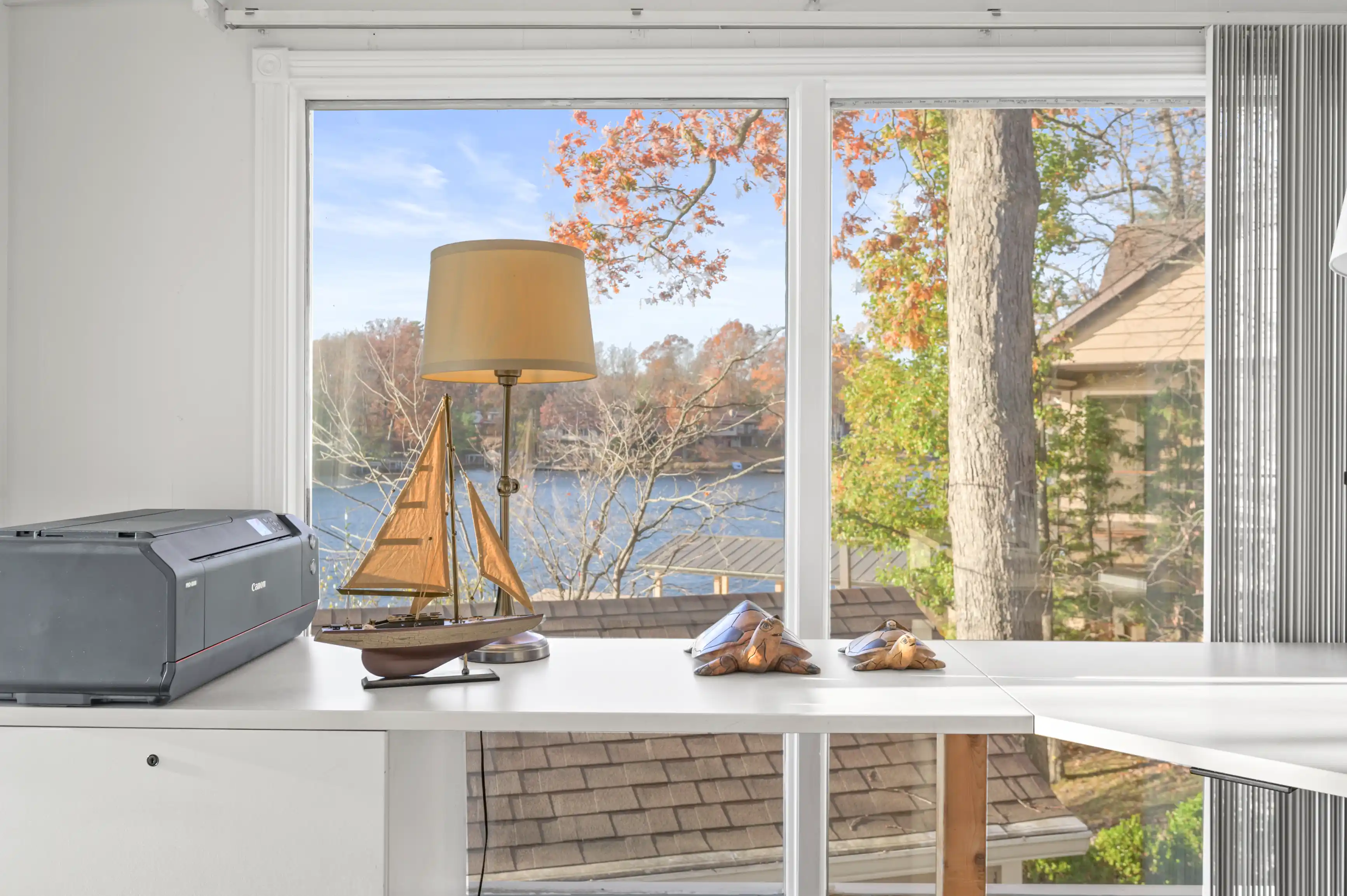 Bright home office with a white desk featuring a printer, decorative sailing ship, and sunglasses, with a lamp and a window overlooking a lake surrounded by autumn trees.