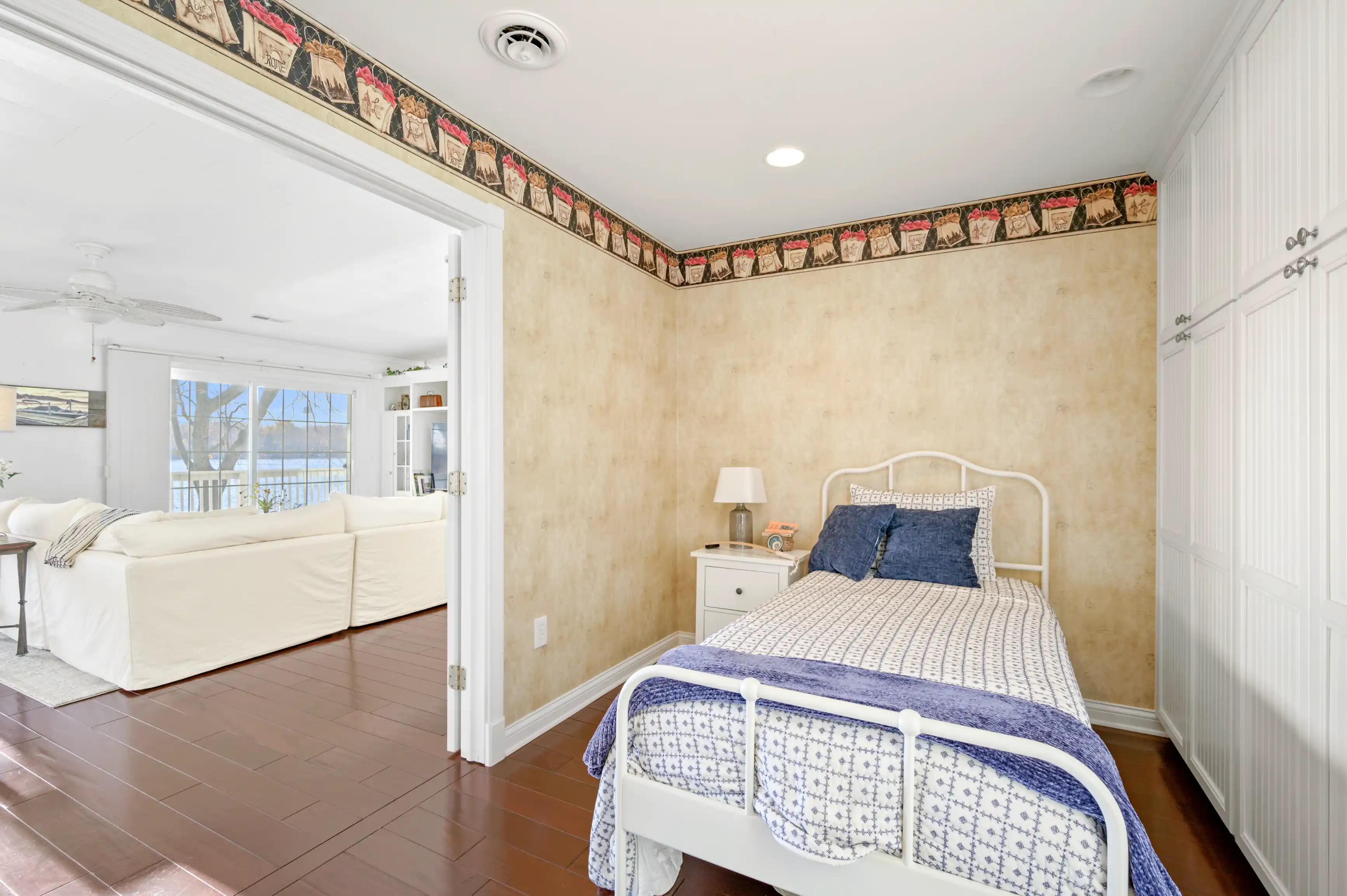 Bright and welcoming bedroom interior with a blue and white bedspread, beige walls, and tiled flooring, leading into an adjoining living room area with white sofa and a view of a waterfront through large windows.