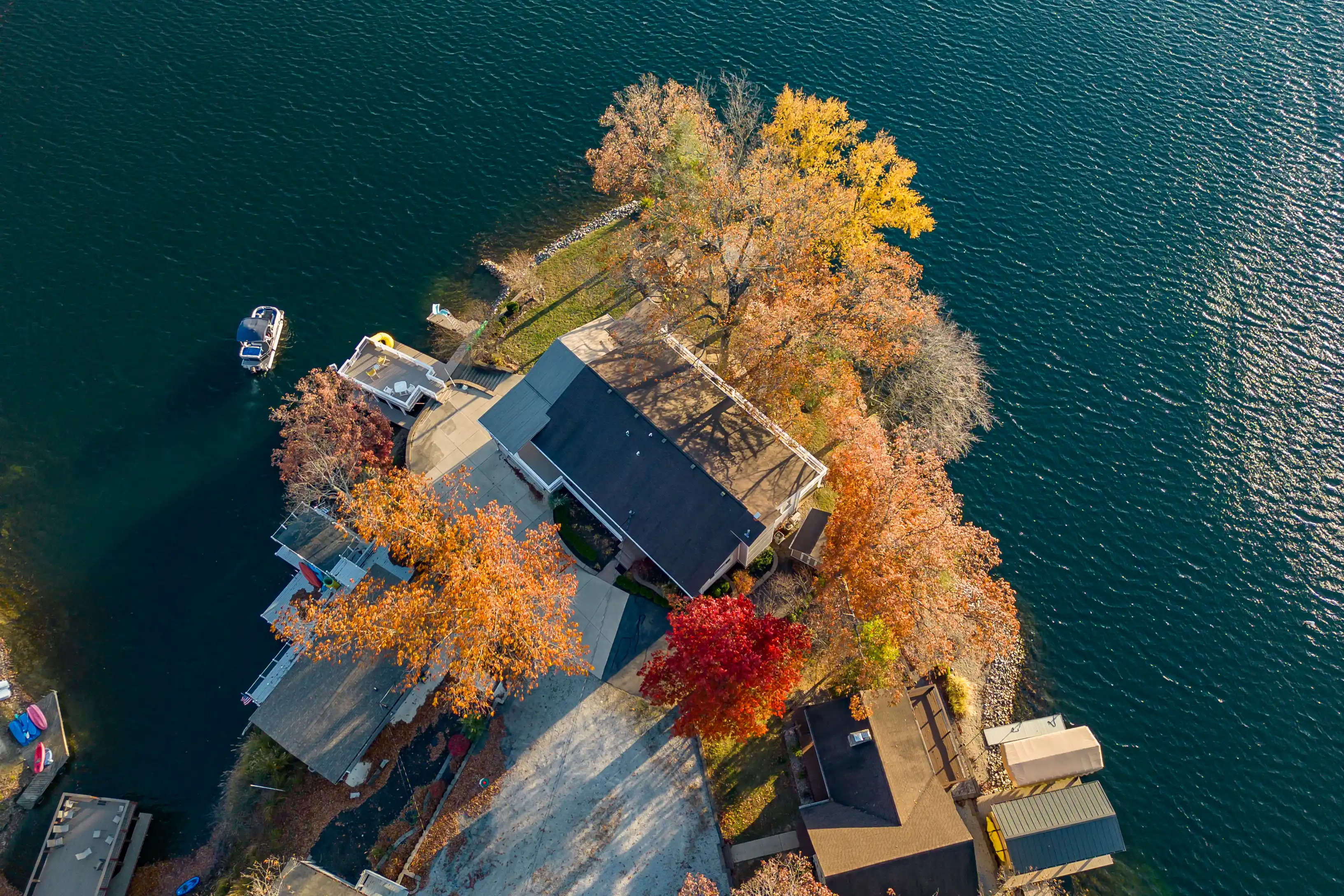 Aerial view of a lakeside home surrounded by autumn-colored trees, with a boat docked at a pier nearby.