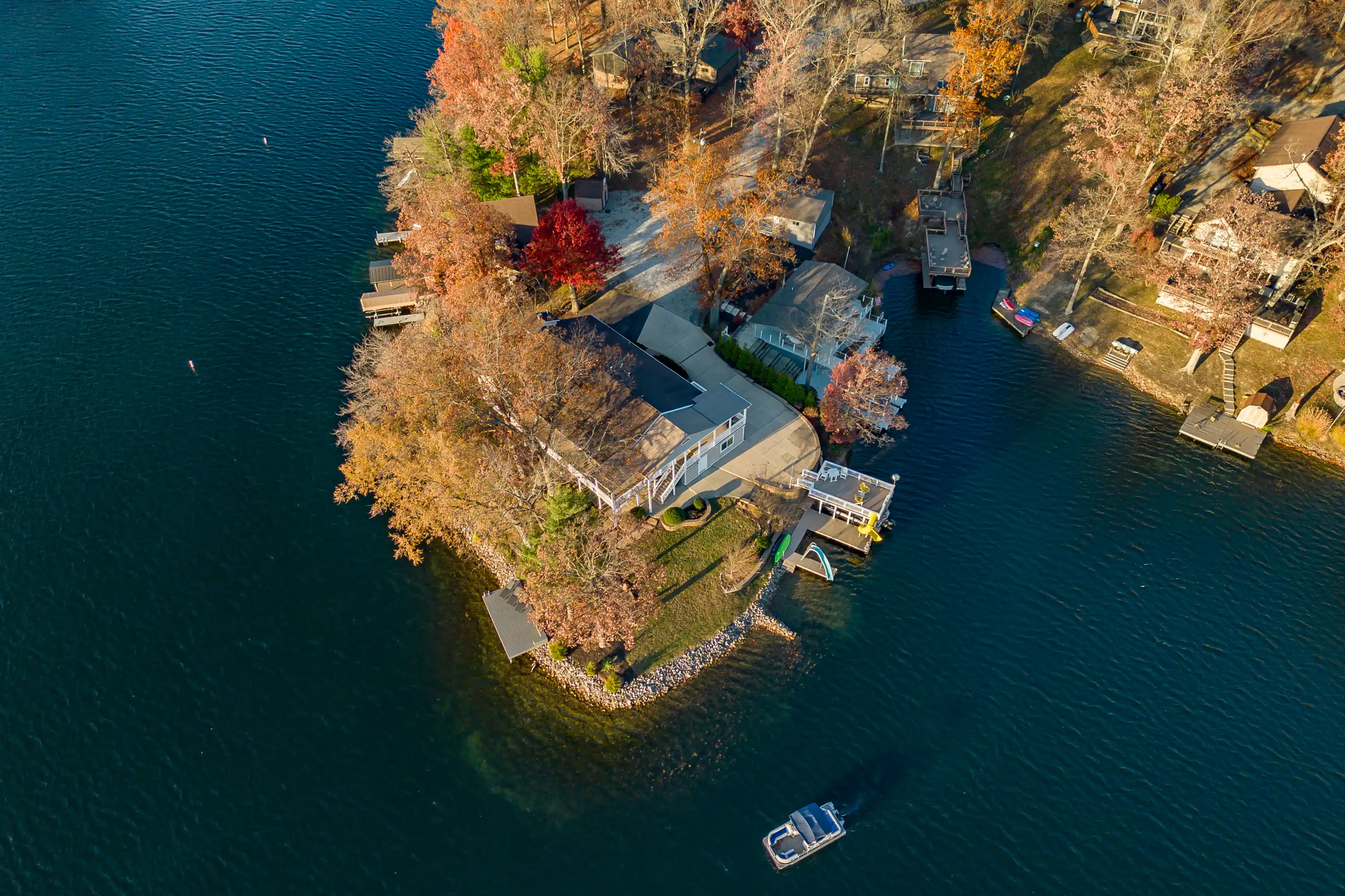 Aerial view of a lakeside residential area with autumnal trees and docks, with a boat cruising on the water.