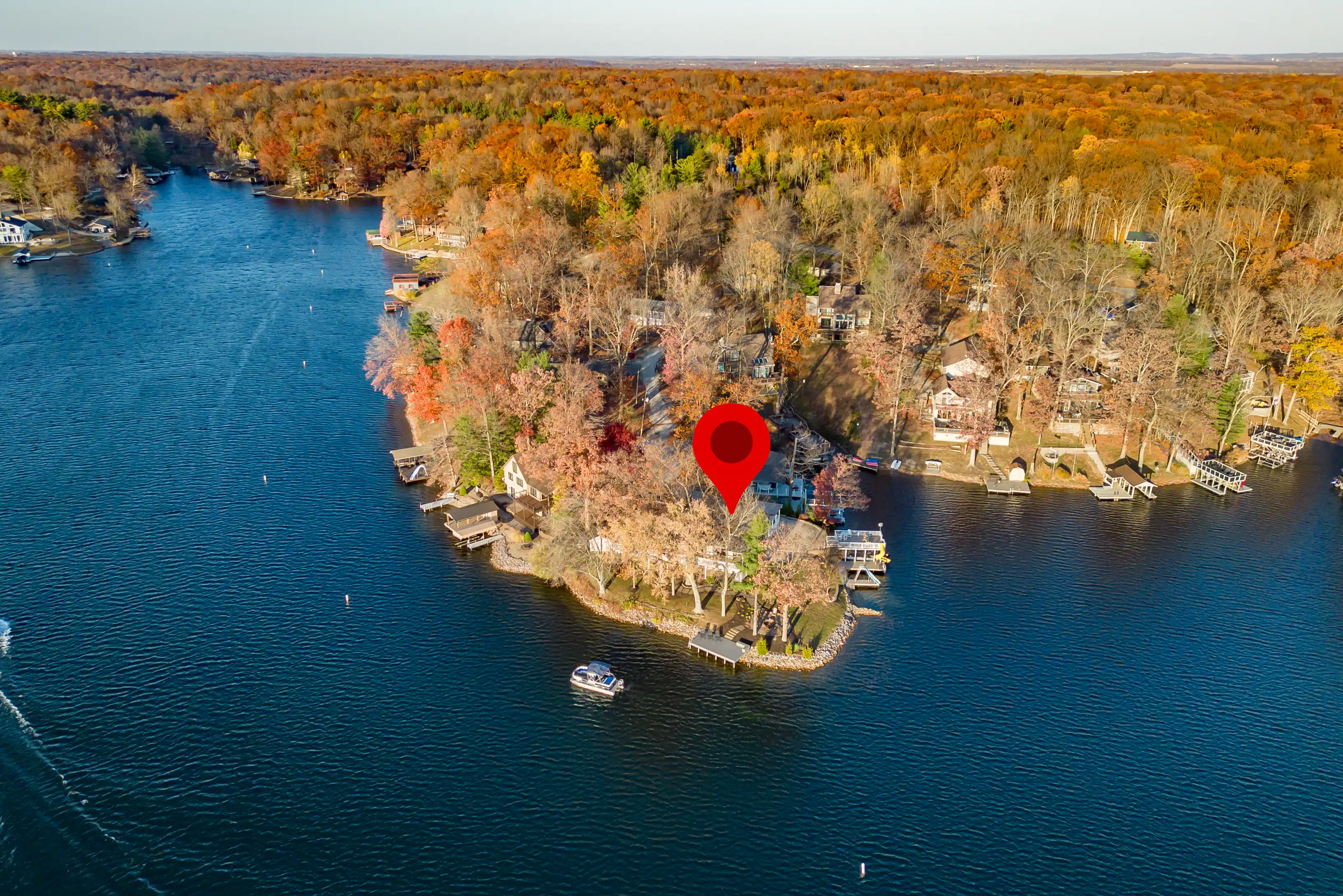 Aerial view of a lake with a peninsula dotted with houses amid colorful autumn trees, marked by a red location pin.
