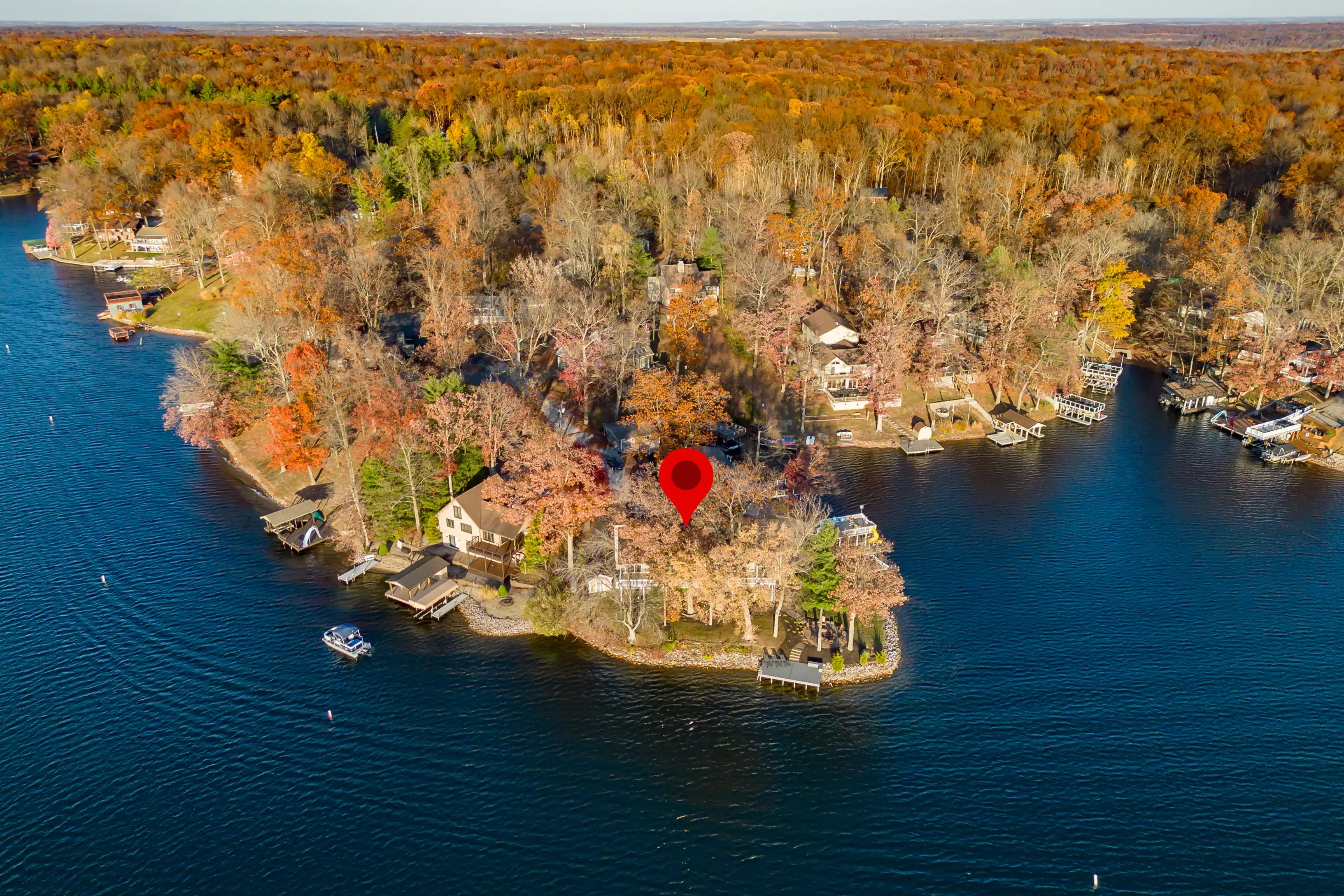 Aerial view of a lake with a red location marker over a peninsula with houses surrounded by autumn-colored trees.