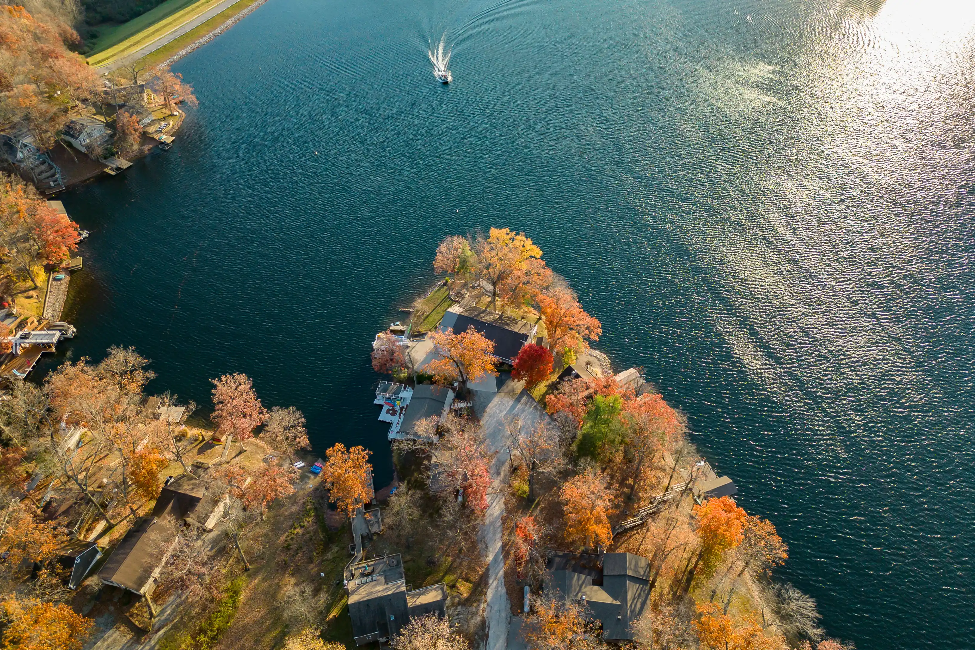 Aerial view of a forested peninsula with autumn foliage extending into a large lake, with boats and docks along the shoreline and a small speedboat creating ripples on the water.