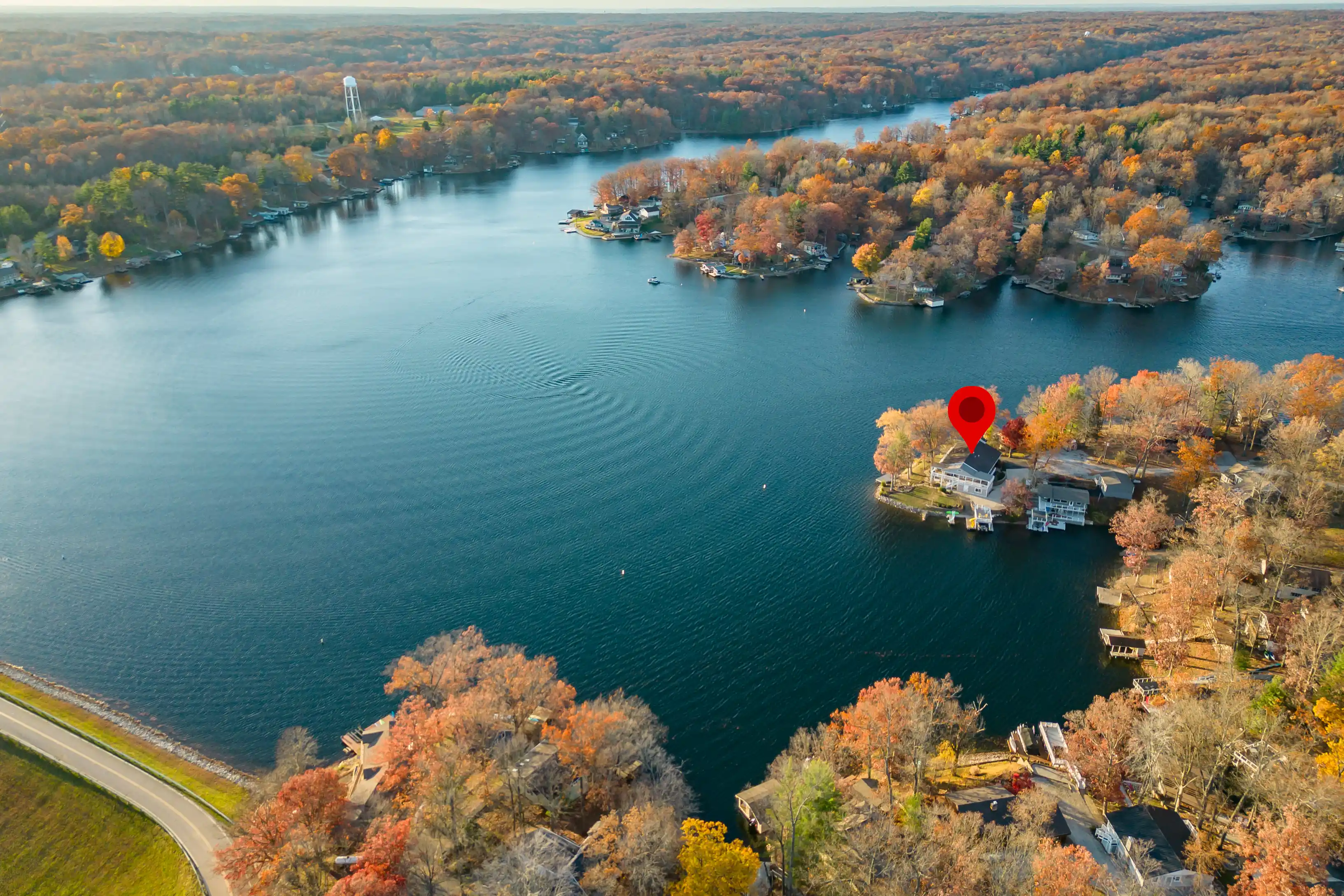 Aerial view of a lake surrounded by houses with docks amidst autumn-colored trees, with a red location marker pinpointing a specific property.