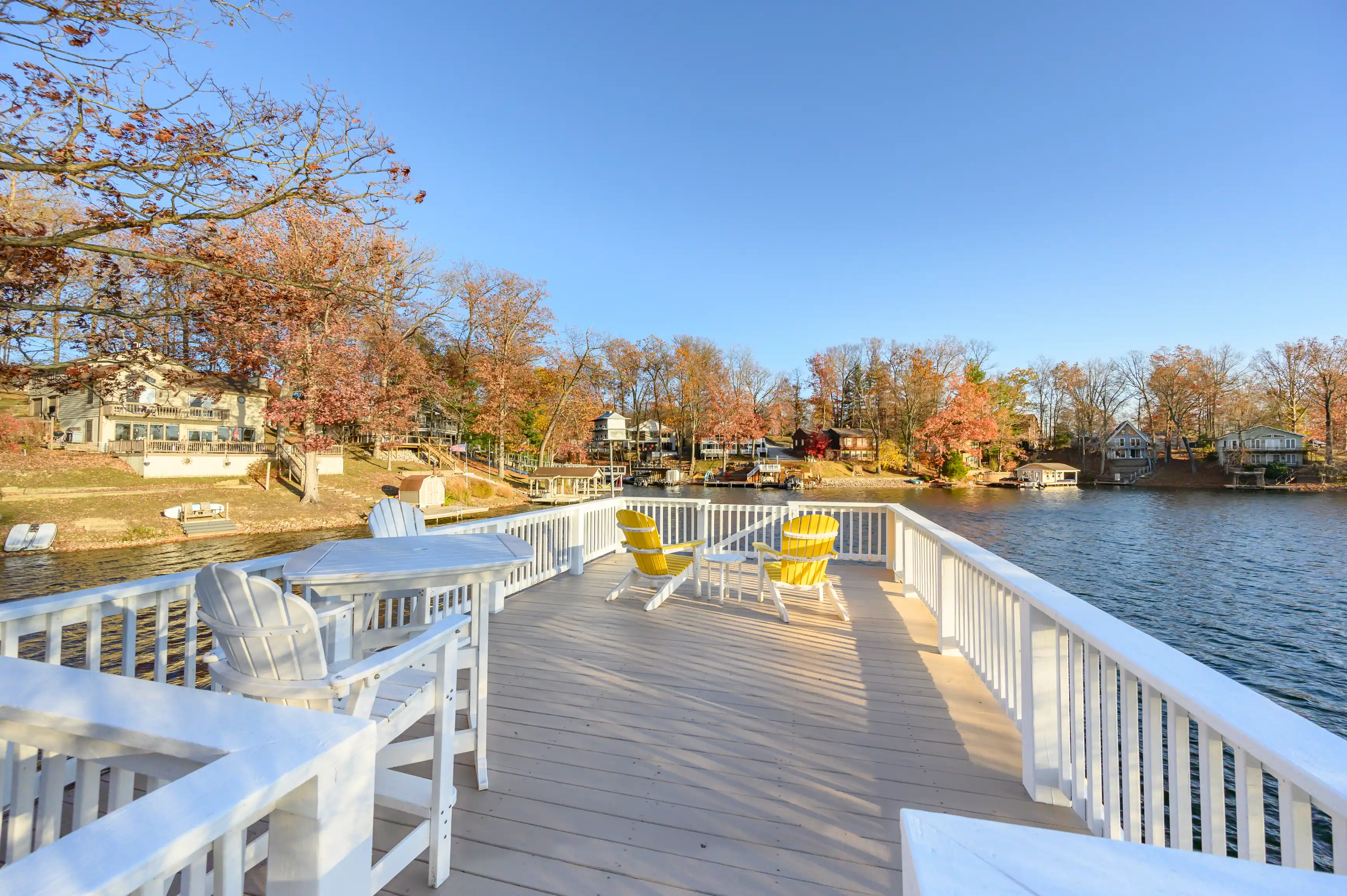 Alt text: A spacious wooden deck with white railing overlooking a tranquil lake with autumn trees and houses along the shore, featuring several Adirondack chairs for relaxation.