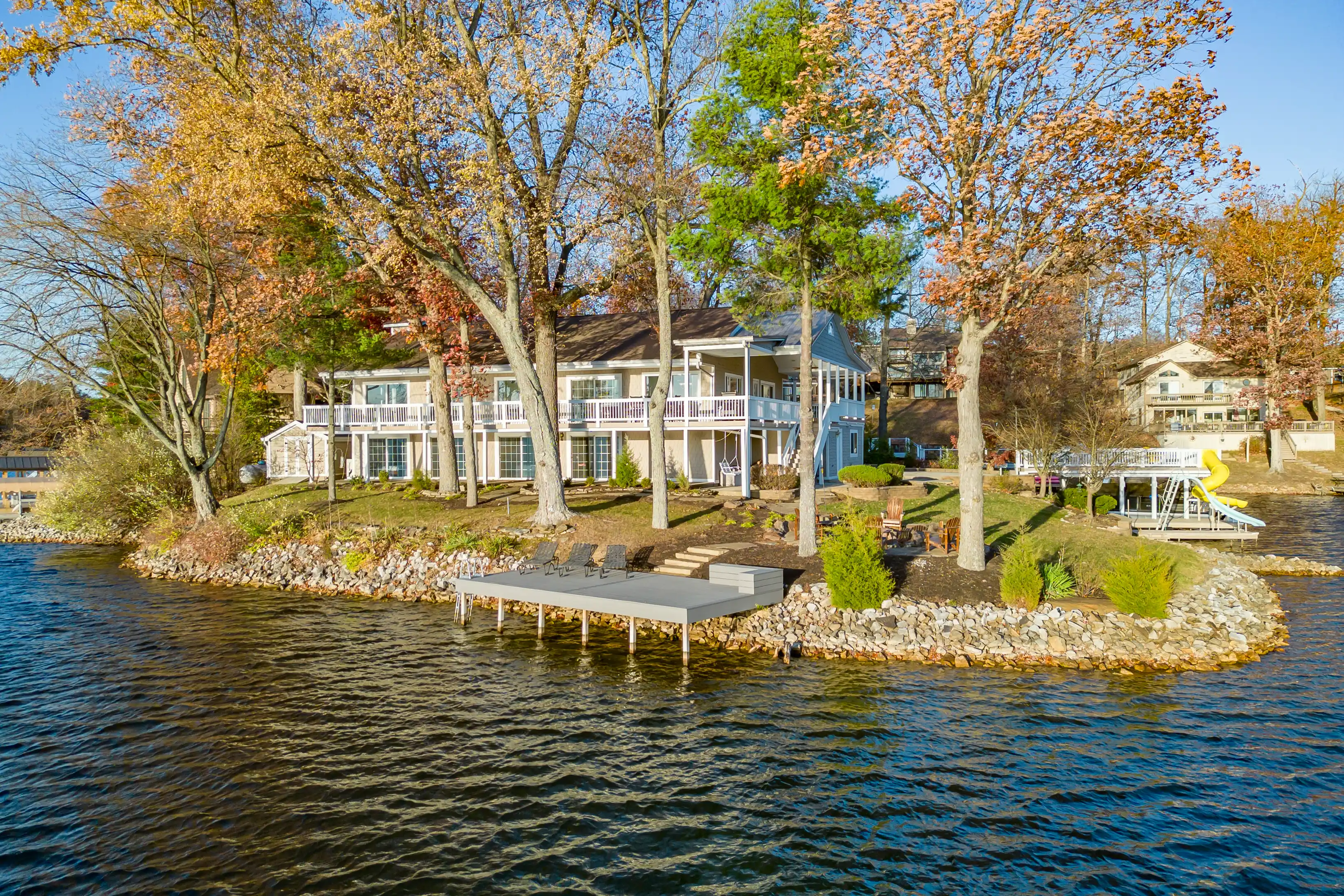 Lakefront homes with porches and autumn trees, featuring a small pier and a yellow slide into the water.