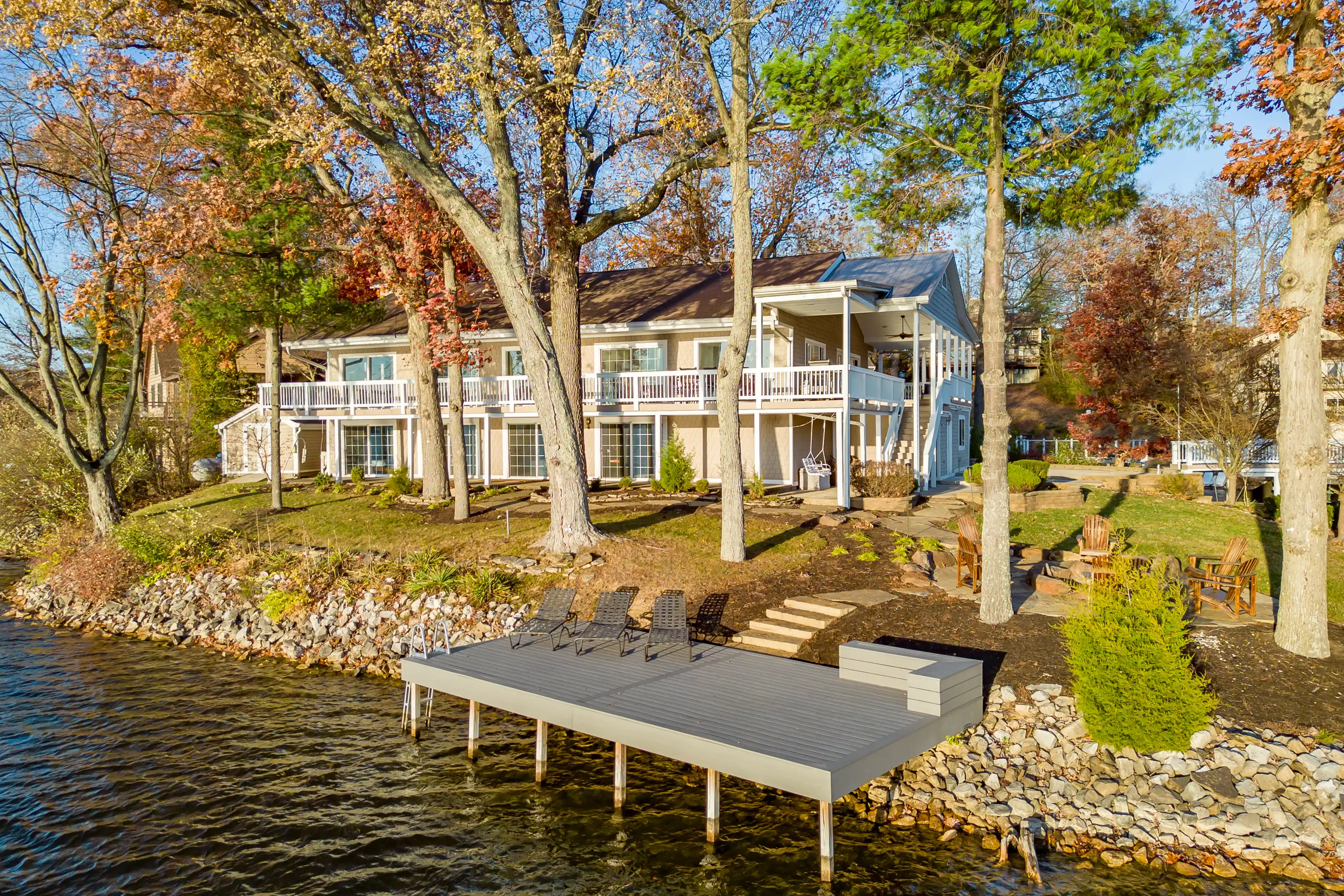 Lakeside two-story houses with balconies surrounded by autumn trees, a wooden dock with chairs on the foreground.