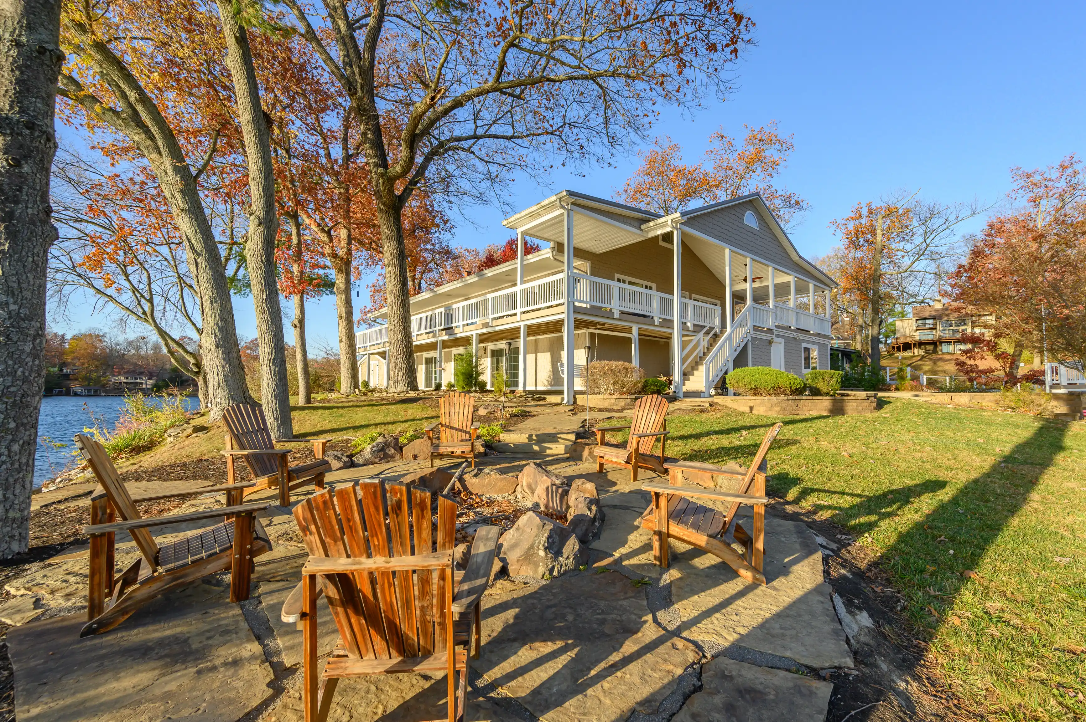 Wooden chairs arranged around a fire pit by a lakefront house with a porch during autumn.