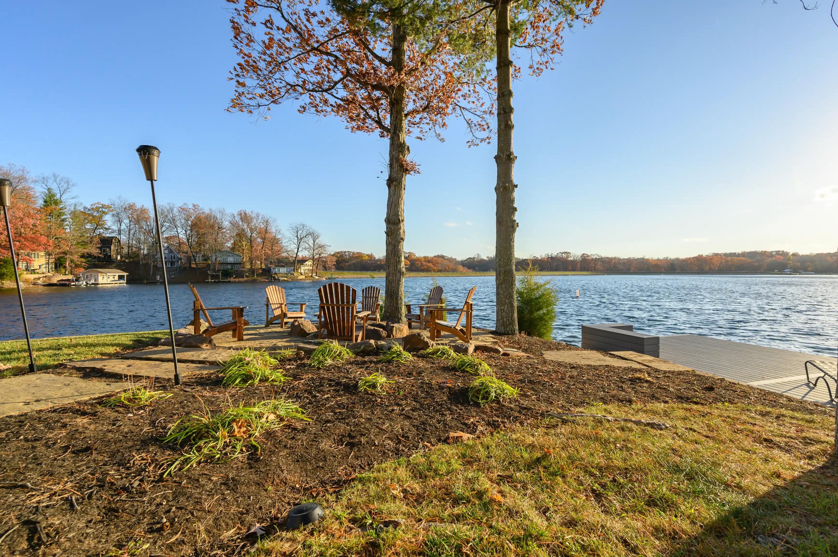 Scenic view of a lakeside with Adirondack chairs on the shore, trees with autumn leaves, and houses across the water under a clear blue sky.