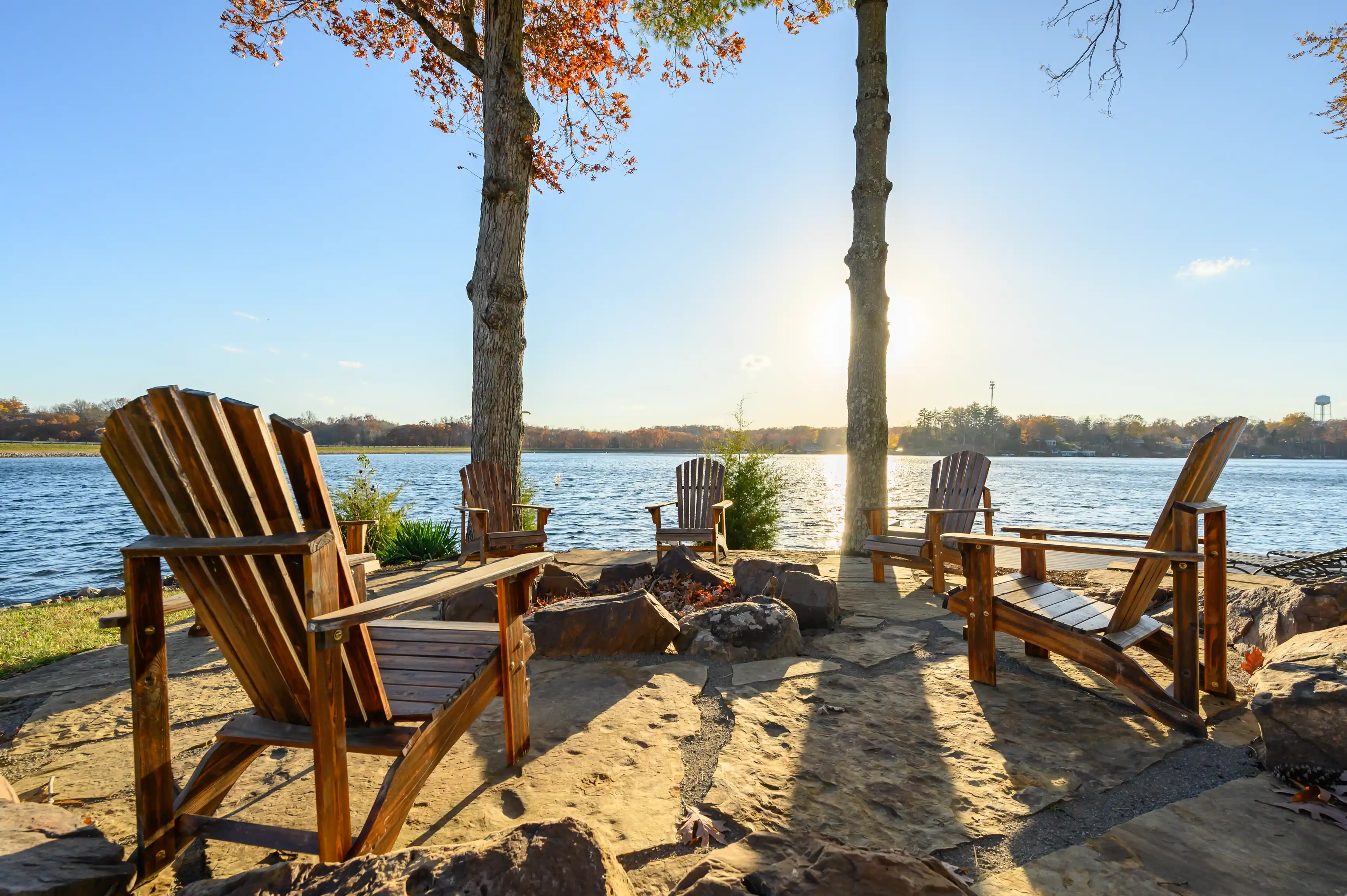 Two wooden Adirondack chairs facing a tranquil lake with a sunset in the background, surrounded by trees with autumn leaves.
