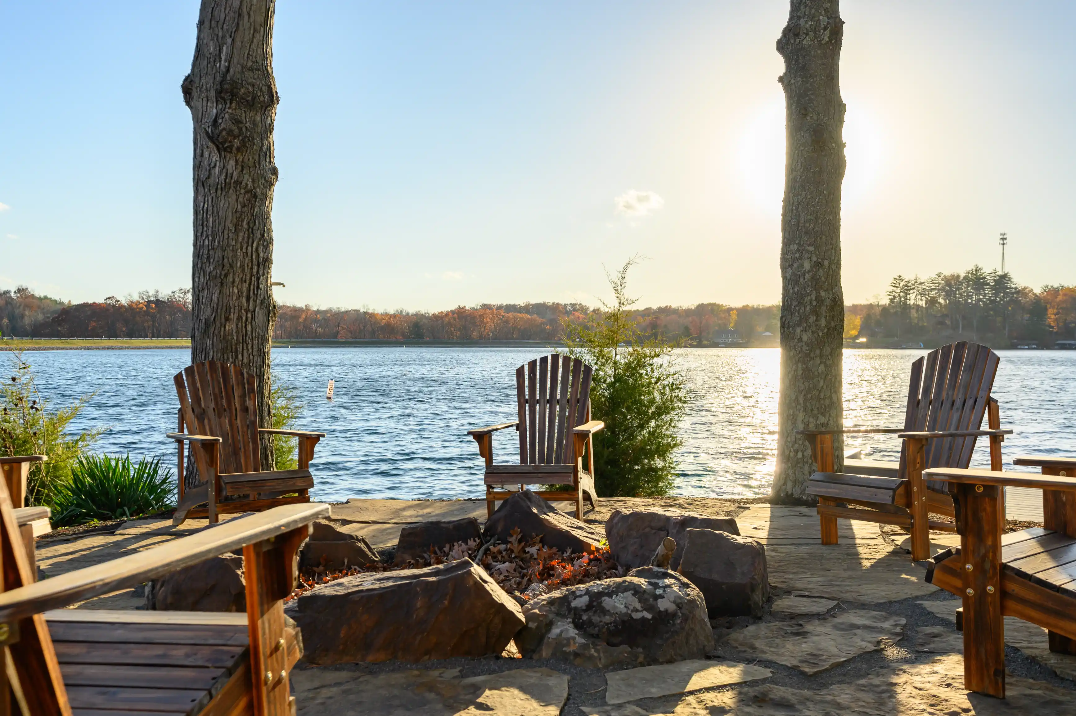 Two wooden Adirondack chairs facing a tranquil lake with a fire pit in the foreground, surrounded by rocks and fallen leaves on a sunny day.