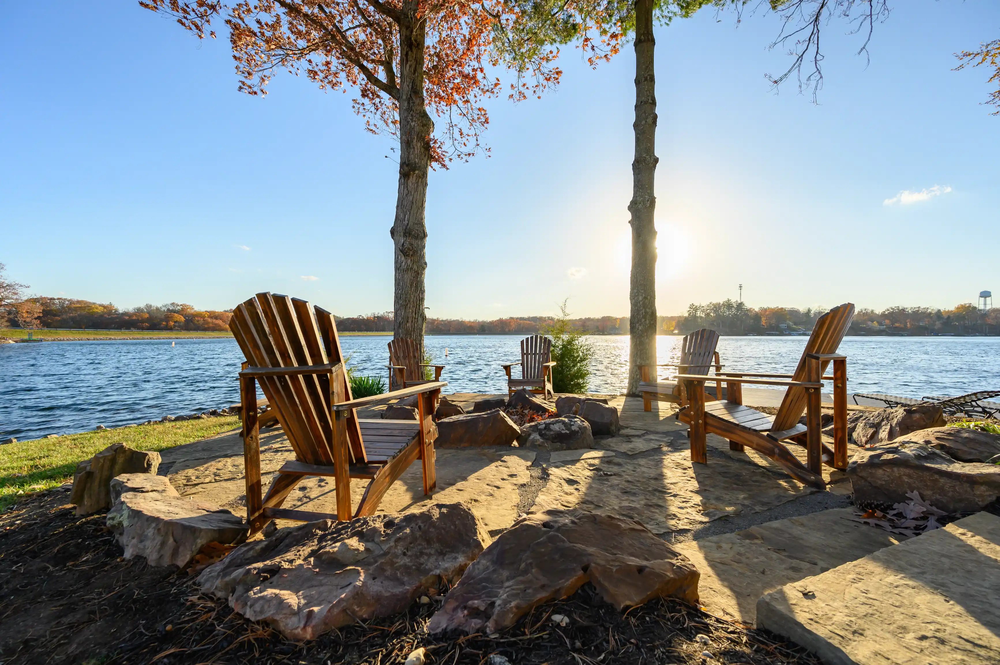 Three wooden Adirondack chairs facing a tranquil lake with a warm sunset backlight, surrounded by autumn colored trees and stone firepit.