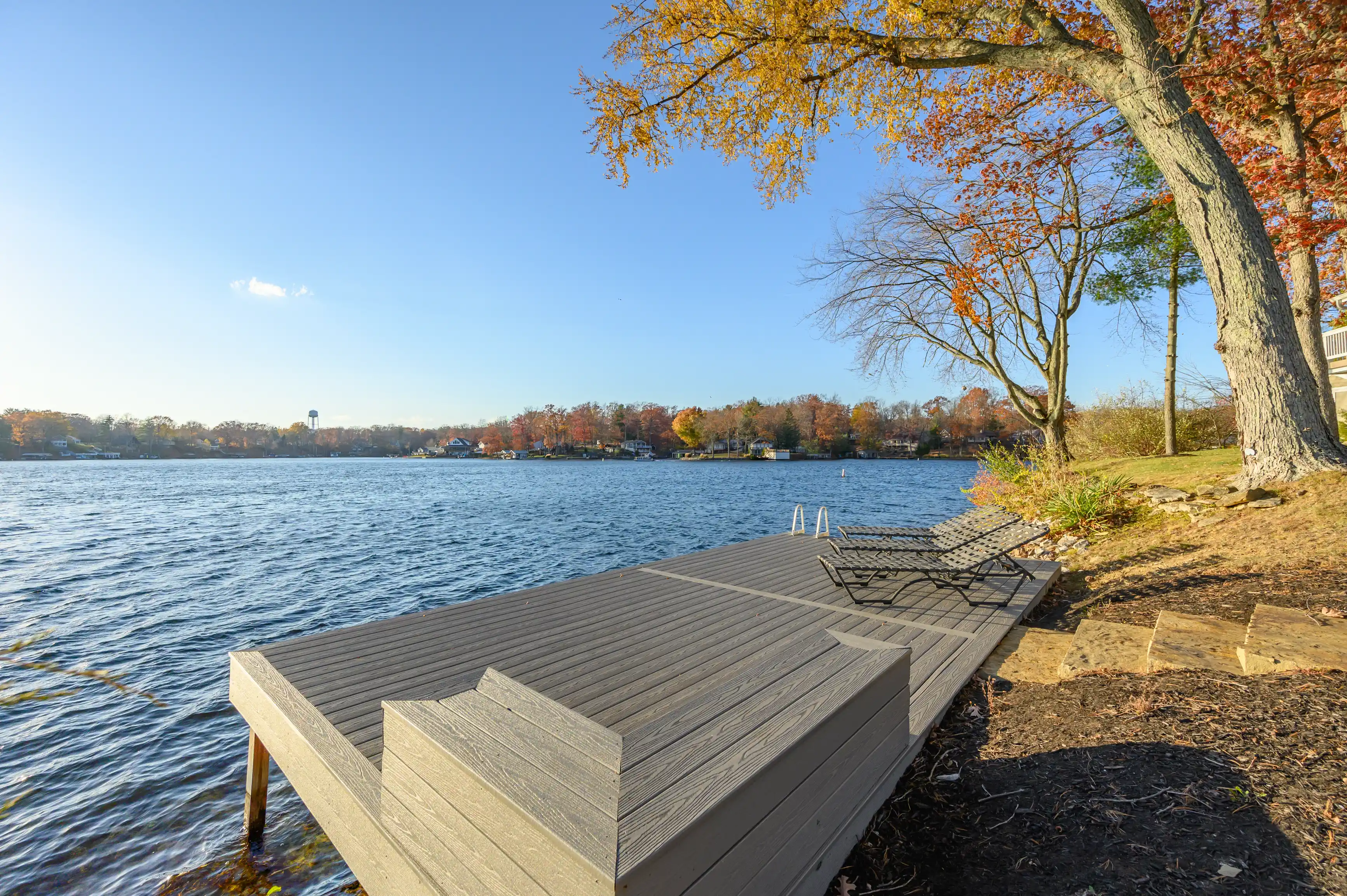 Autumn scene with a wooden dock extending into a peaceful lake, surrounded by trees with colorful fall foliage under a clear blue sky.