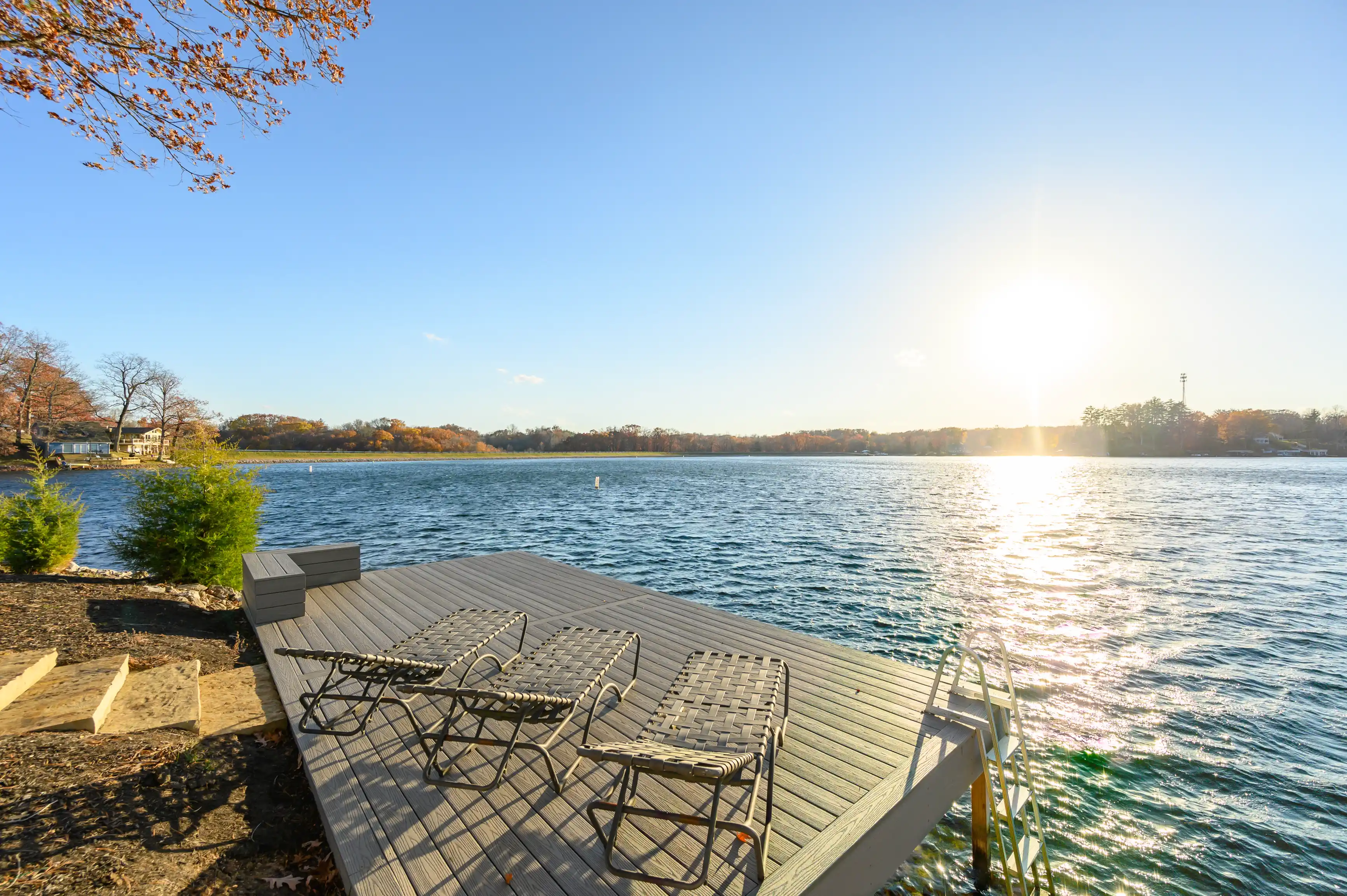 Lake view from a wooden pier with lounge chairs during autumn, with the sun setting over the water creating a sparkling effect.