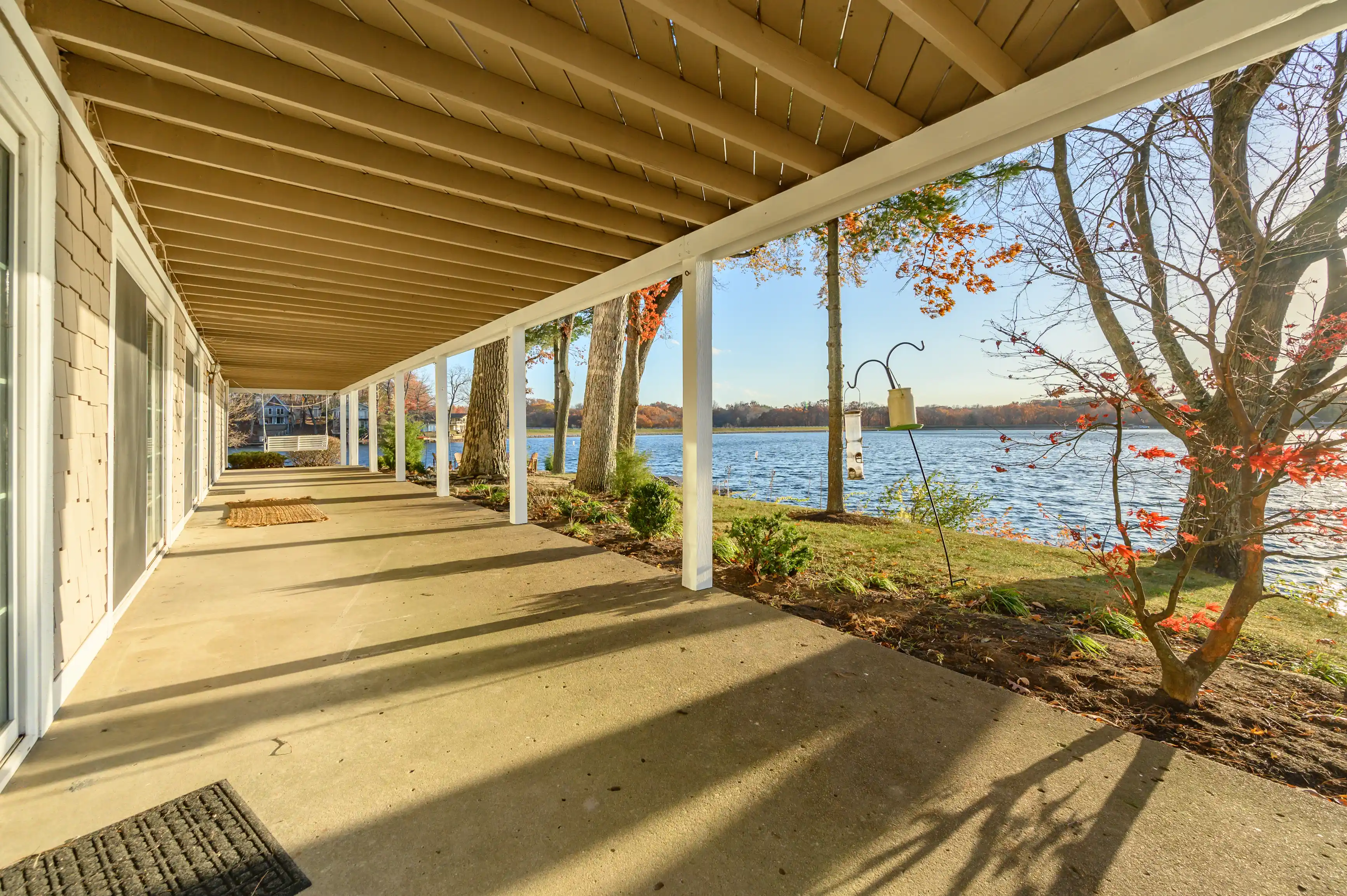 Covered porch of a lakeside house with white pillars, leaf-bearing trees, and a clear view of the lake under a sunny sky.