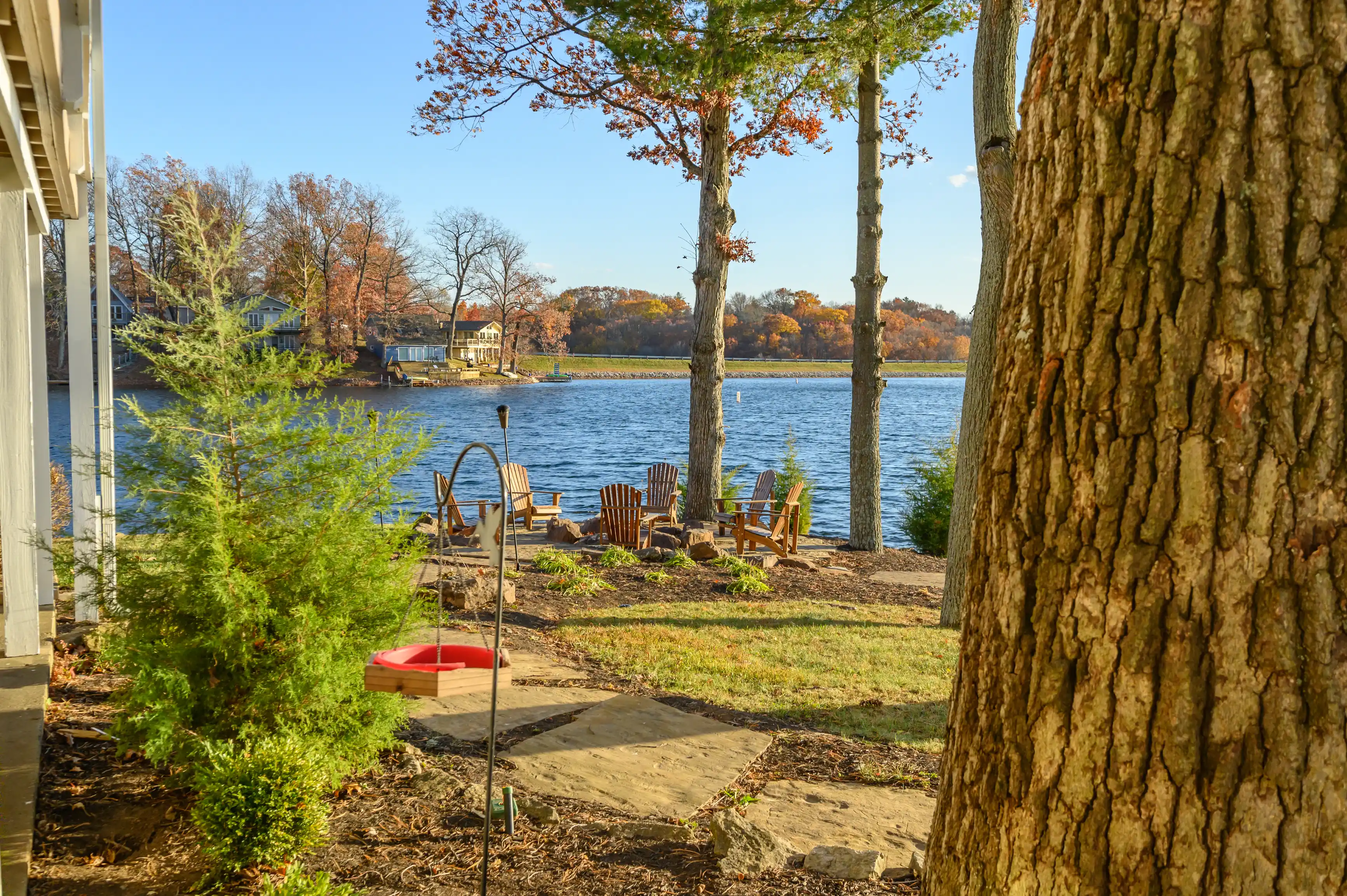 Tranquil lakeside backyard with Adirondack chairs, autumn trees, and a clear blue sky.