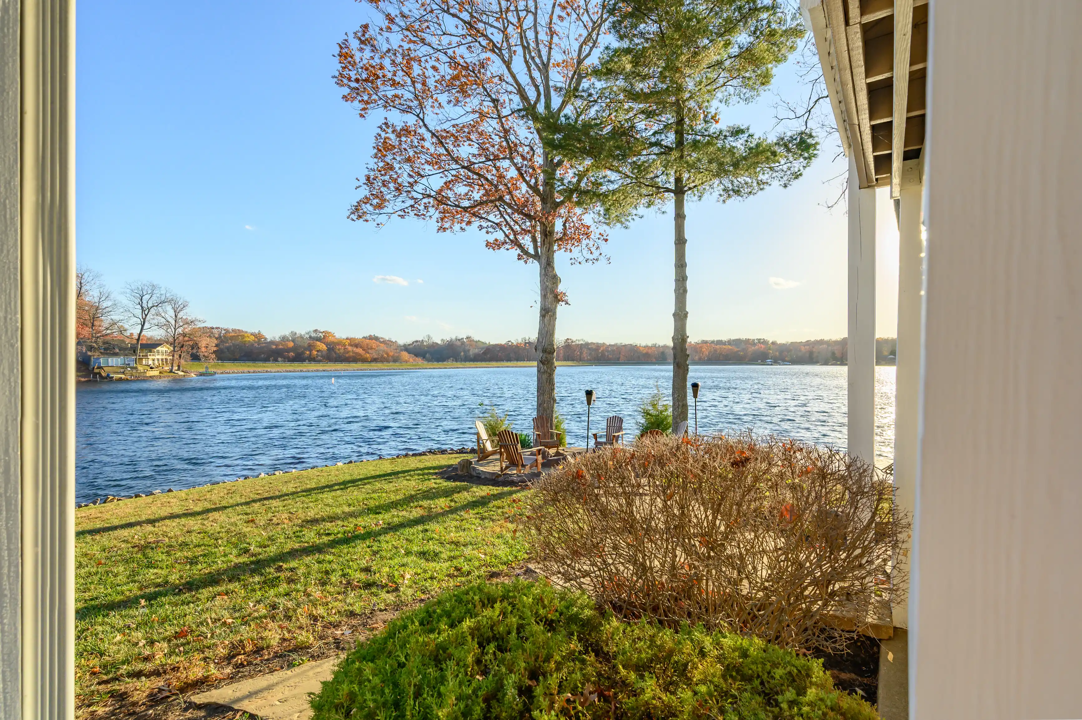 Lake view from a backyard with Adirondack chairs by the water's edge, trees with autumn leaves, and a clear blue sky.