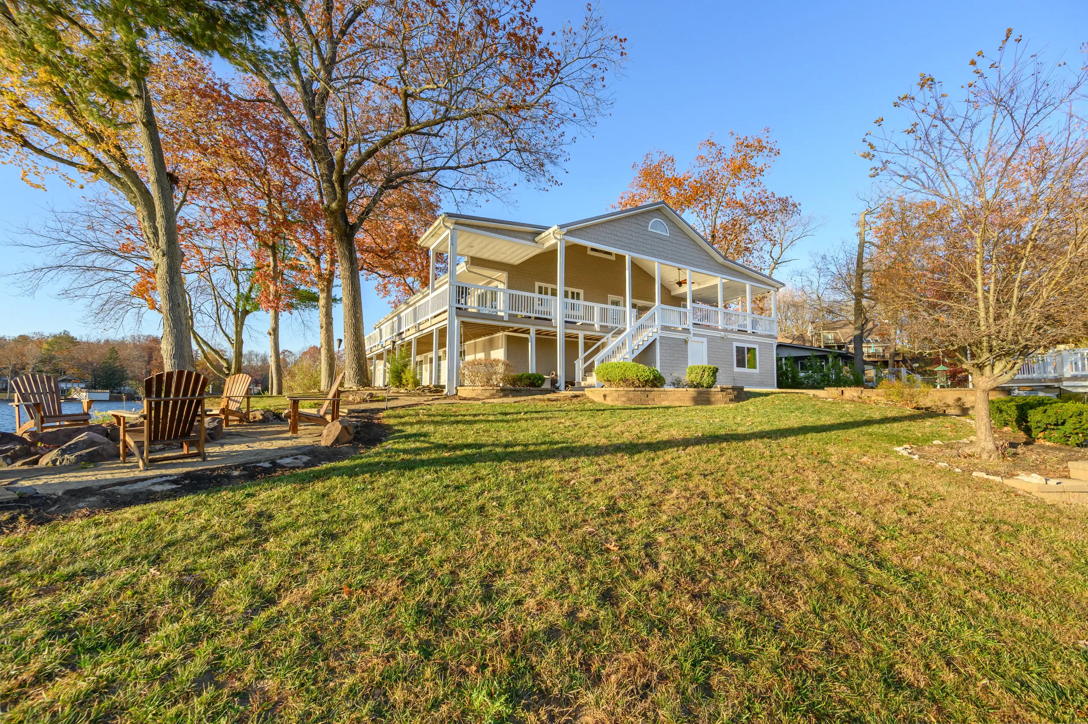 Two-story house with a wrap-around porch surrounded by autumn trees with Adirondack chairs overlooking a lake.