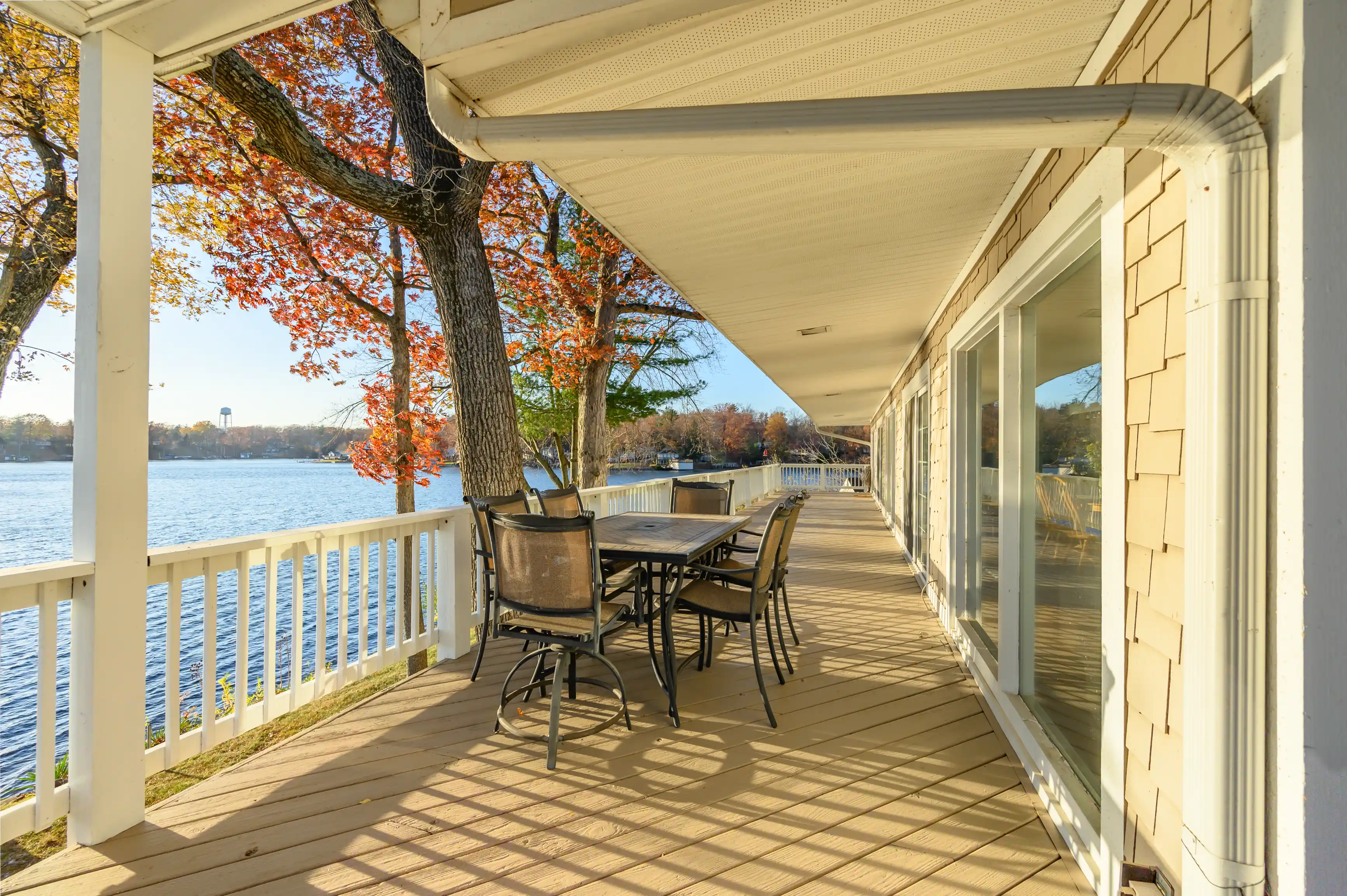 Spacious lakefront porch with outdoor dining furniture set, overlooking a tranquil lake with autumn foliage on a clear sunny day.