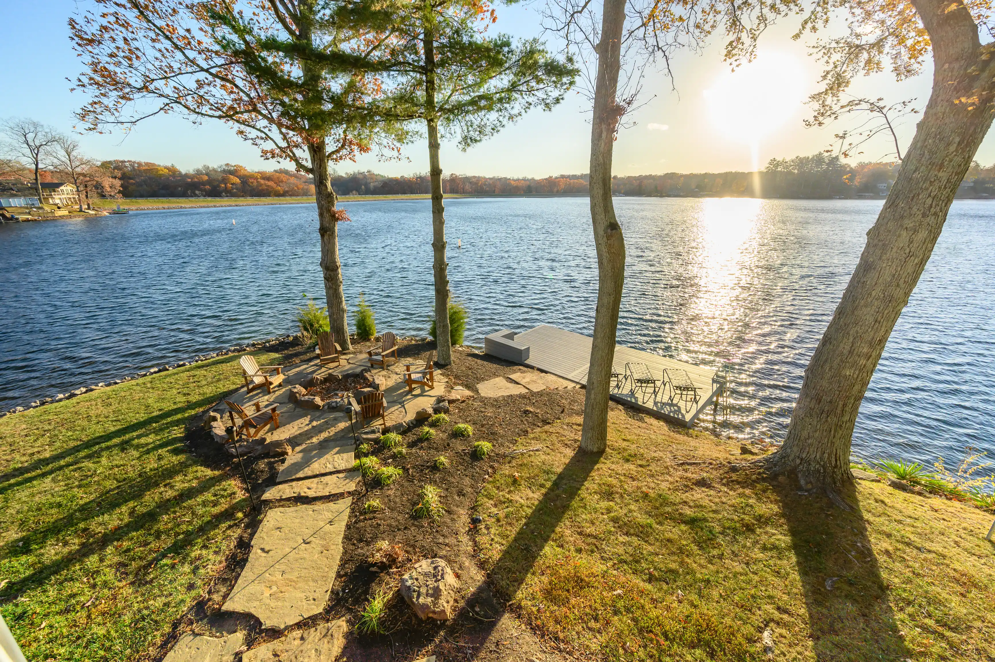 Lakeside backyard with Adirondack chairs, a fire pit, and a wooden pier extending into a tranquil lake surrounded by trees with autumn foliage under a sunny sky.
