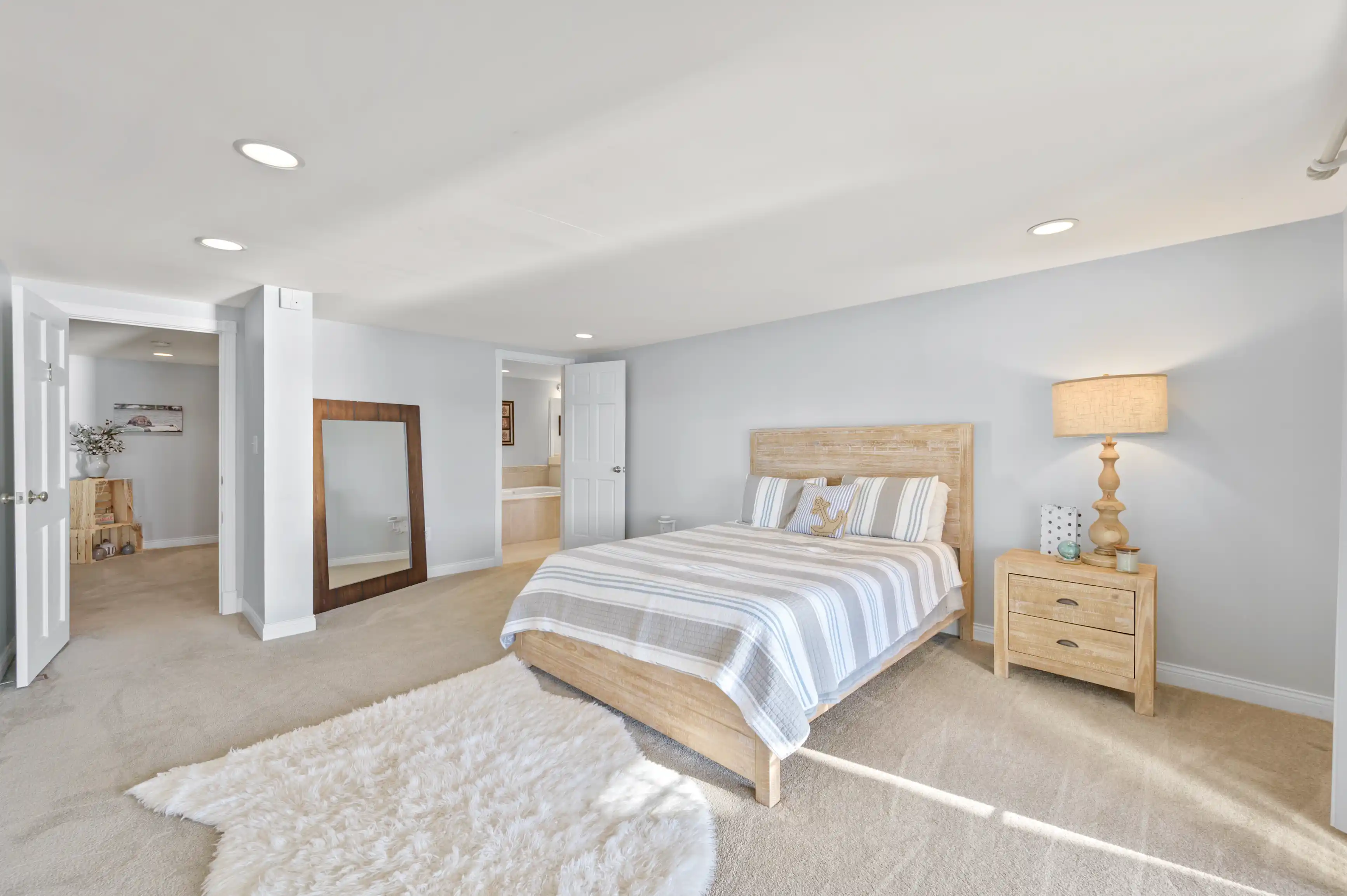 A bright, modern bedroom with a wooden bed and matching side table, striped bedding, a large floor mirror, a white fluffy area rug, and an en suite bathroom entrance visible in the background.