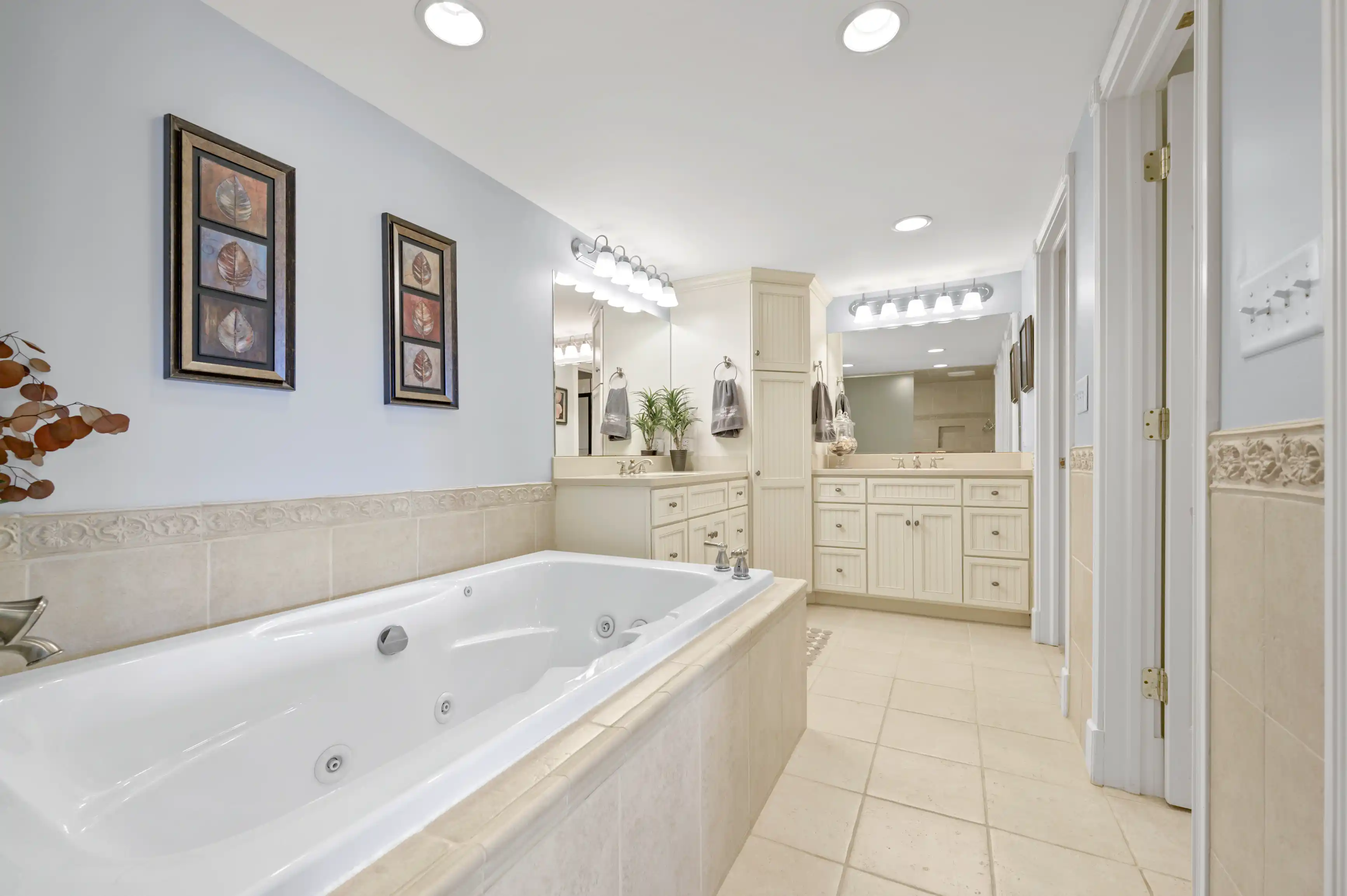 Bright and spacious bathroom with a large jacuzzi tub, double vanity sink with a large mirror and vanity lights, framed artwork on the walls, and tiled flooring.